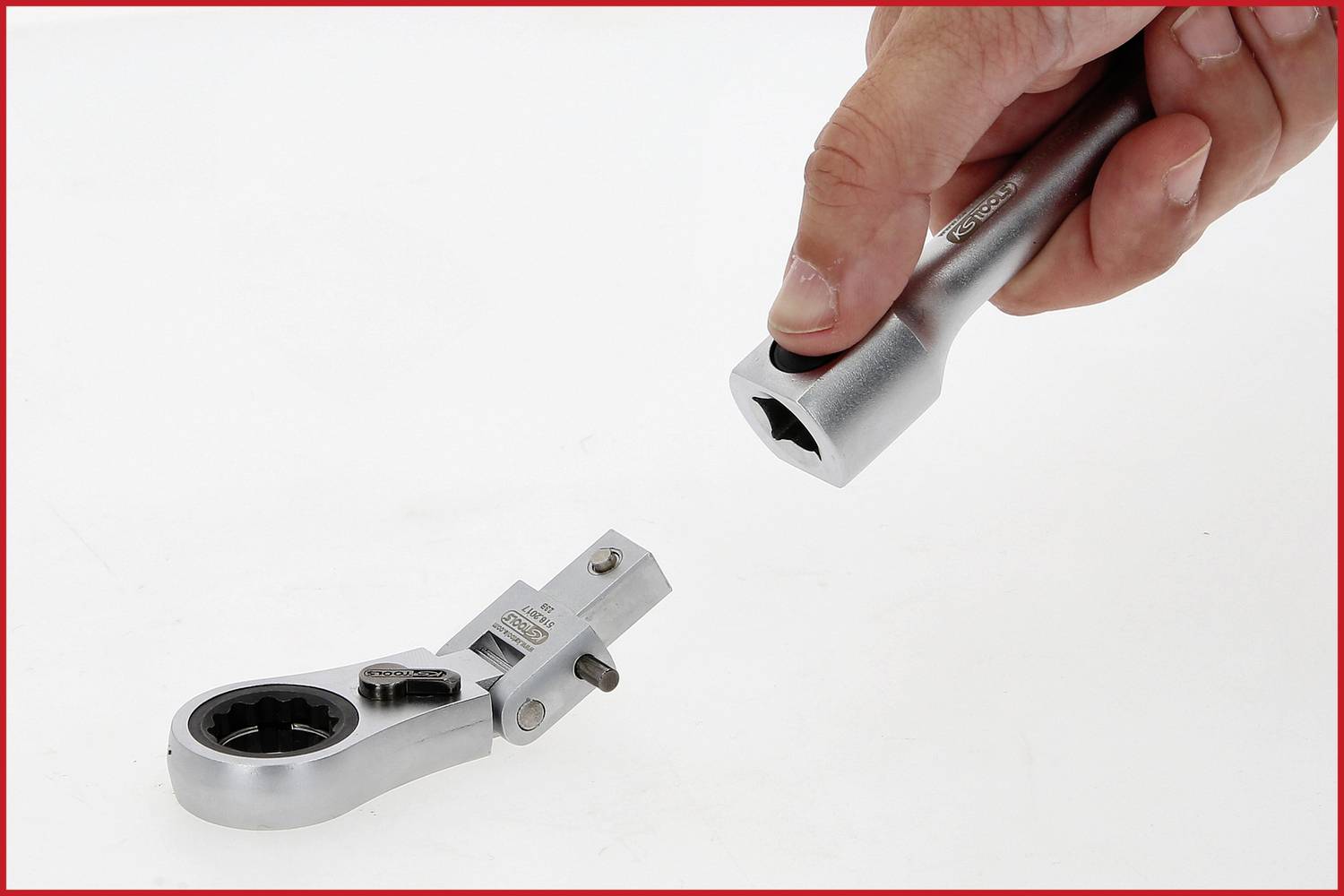 Hand hält Steckschlüssel in der Nähe eines Ratschenkopfes, beide aus Metall. Sie sind getrennt und auf einer weißen Oberfläche platziert.