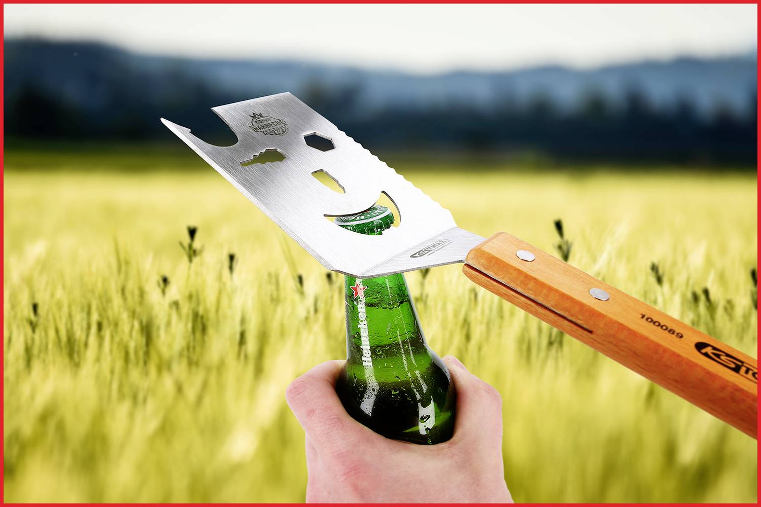 Ein Flaschenöffner in Form einer Handsäge öffnet eine Bierflasche vor einem Weizenfeld, im Hintergrund verschwommene Berge.