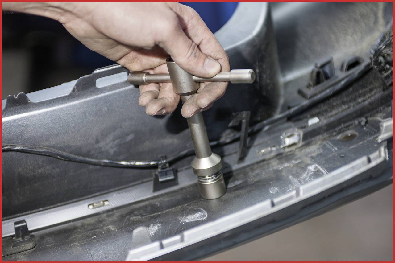 Eine Hand benutzt ein Schraubwerkzeug, um ein Metallteil an einem Auto zu reparieren. Staub und Kabel sind im Hintergrund sichtbar.