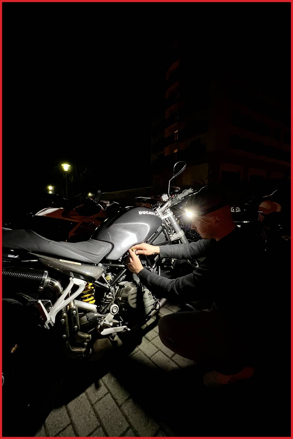 Eine Person arbeitet nachts an einem Motorrad auf einem dunklen Gehweg. Eine Taschenlampe beleuchtet die Szene und das Motorrad.