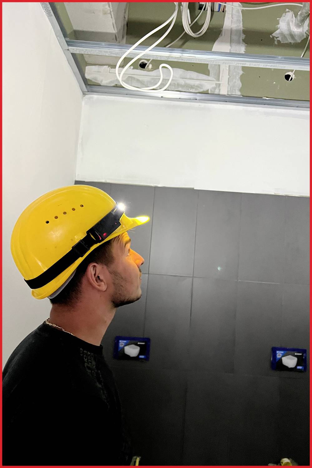 Ein Mann mit gelbem Helm schaut nach oben, während er in einem Raum steht. Es gibt Rohre und Kabel an der Decke.