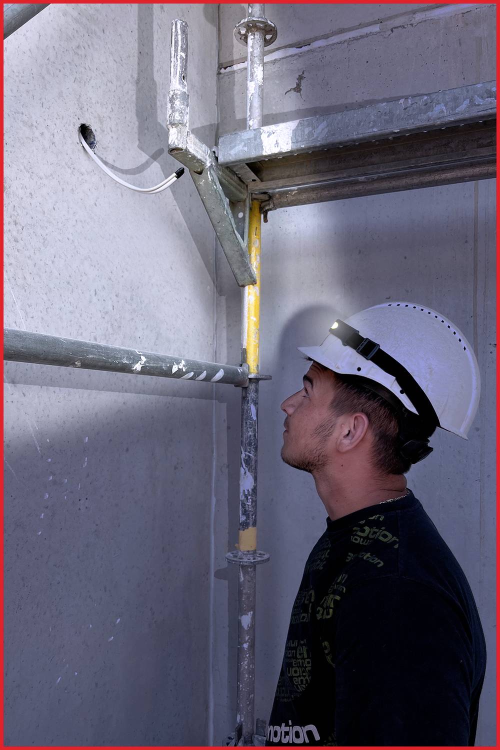 Ein Bauarbeiter mit Schutzhelm betrachtet eine Baustellenwand neben einem Gerüst, während ein Kabel aus einem Loch in der Wand ragt.
