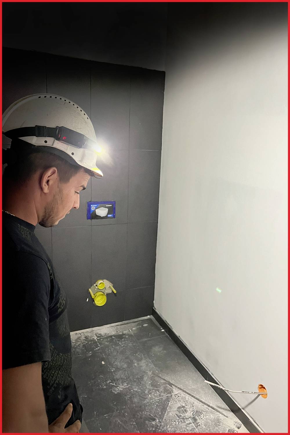 Ein Bauarbeiter mit Helm und Stirnlampe inspiziert eine Baustelle in einem dunklen Raum, neben einer Wand mit verdeckter Steckdose.