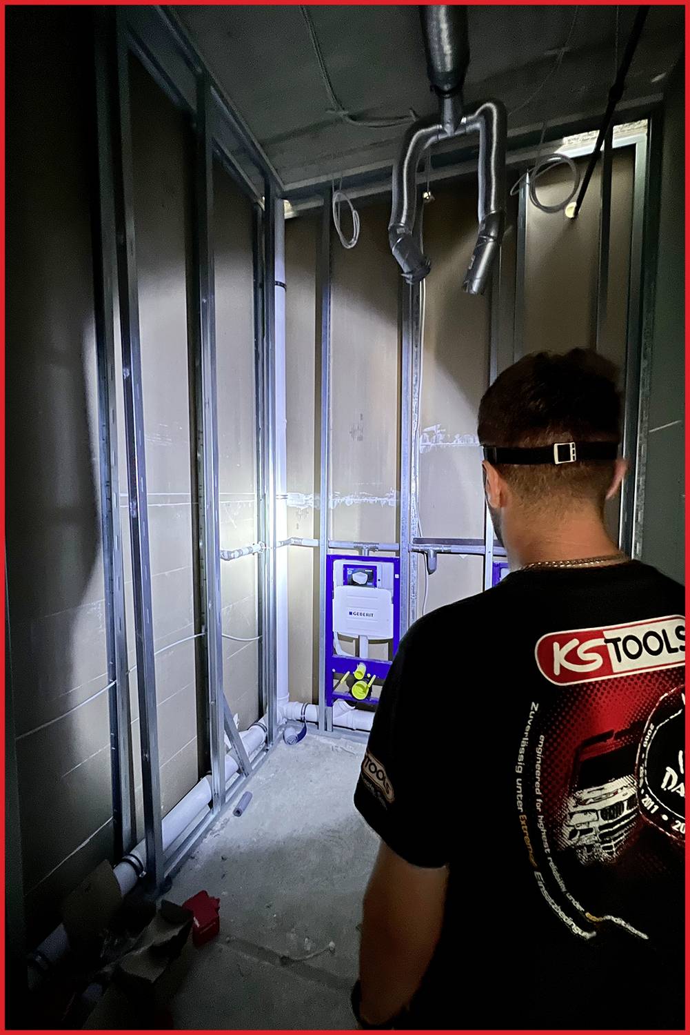 Person, die in einem unfertigen Raum mit sichtbaren Metallrahmen und Rohrleitungen steht. An Wand montierter Spülkasten befindet sich im Hintergrund.