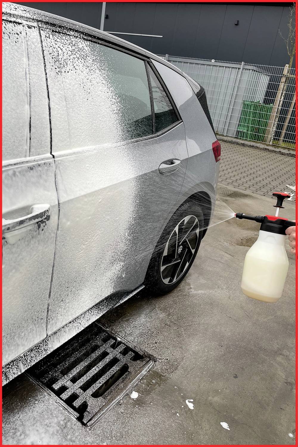Auto wird mit Schaumreiniger besprüht; Fokus auf Heck und Hinterrad. Eine Hand hält eine Schaumspritze. Umgebung: Industriegebiet mit Zaun.