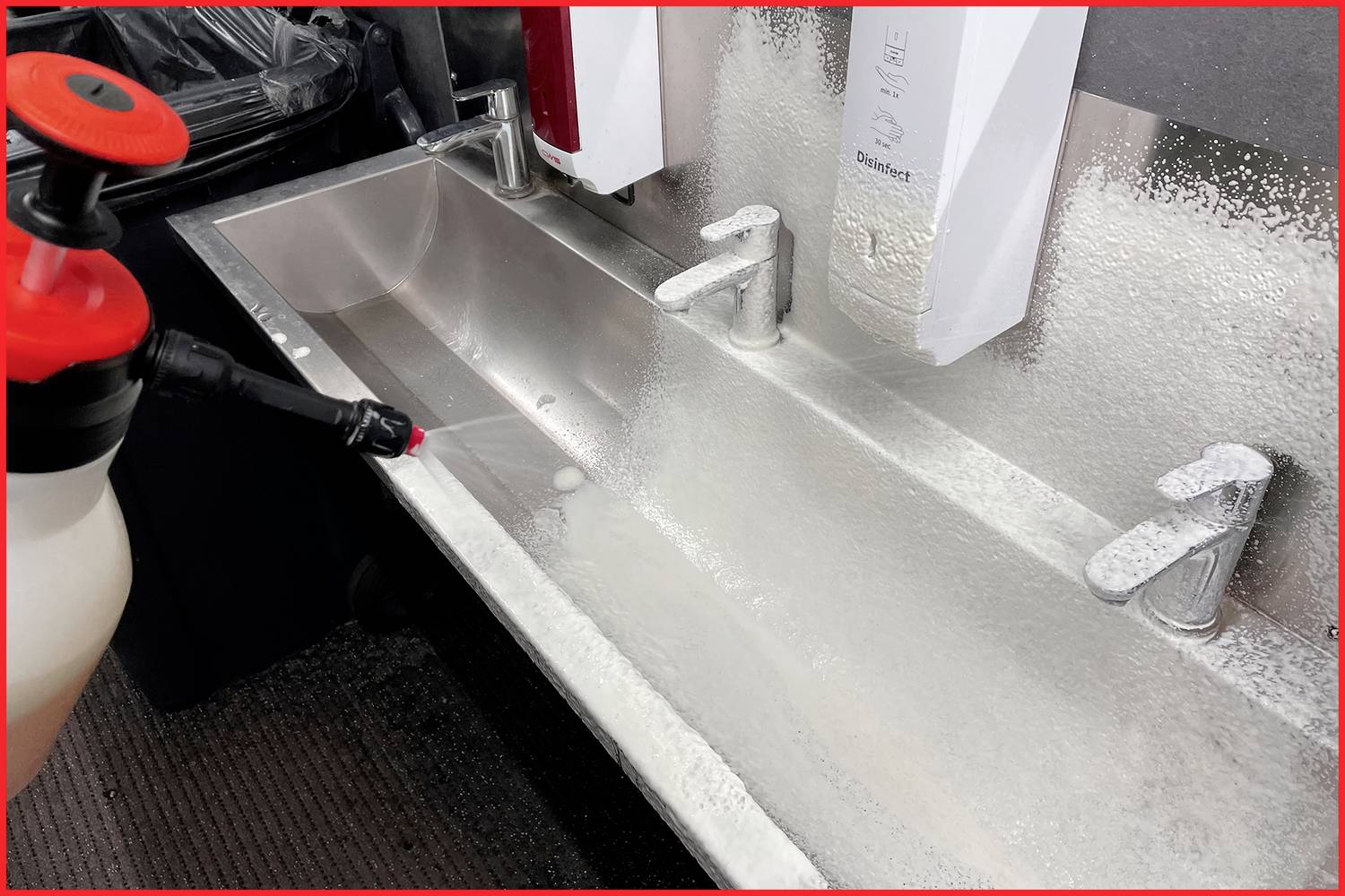 Ein langer Edelstahlschrank mit drei Wasserhähnen zeigt Seifenreste und Schaum, der über die Oberfläche verteilt ist. Ein Reinigungsmittel wird aufgetragen.