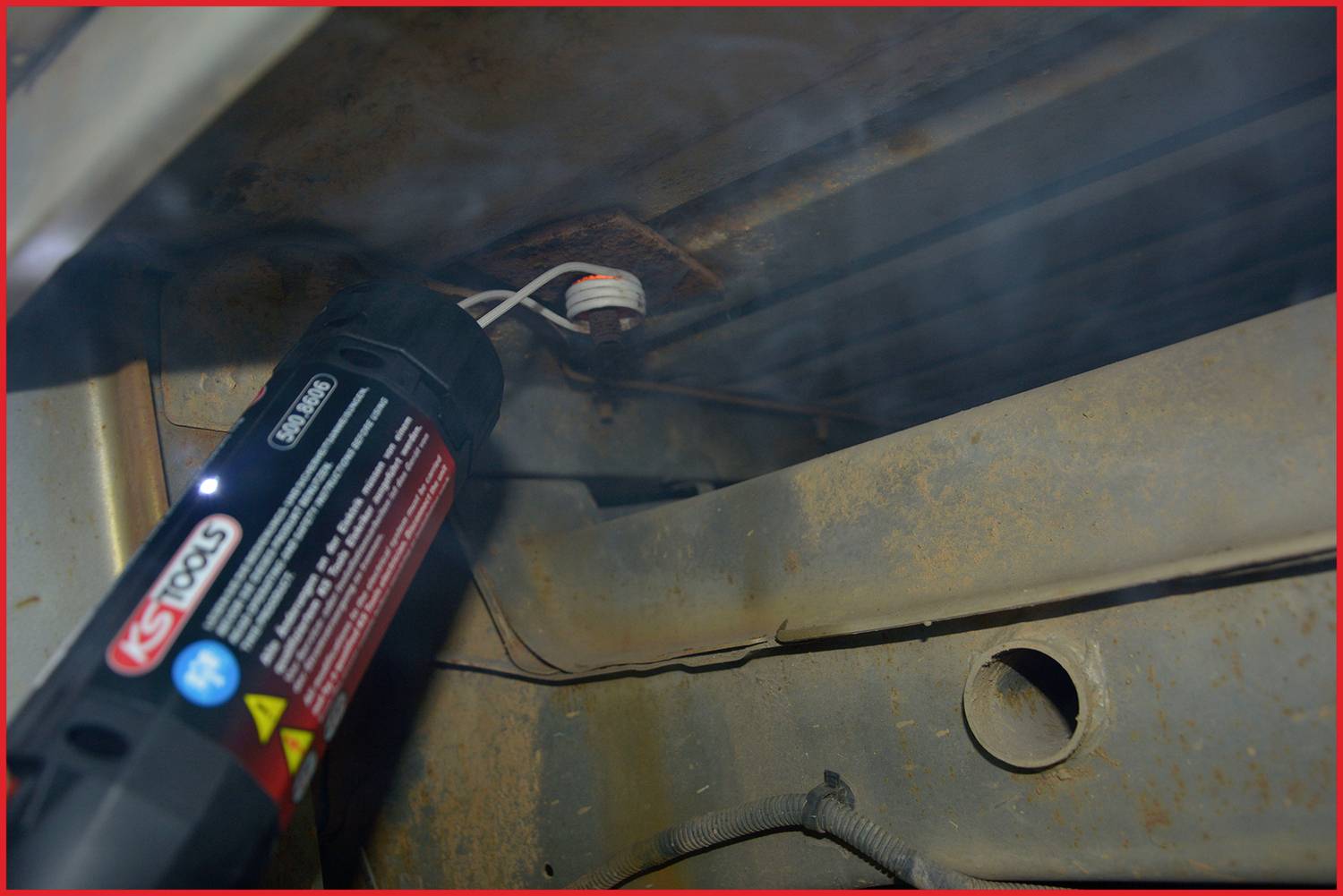 Unterseite eines Autos mit einem Dichtungsmittel-Spray, das auf eine rostige Stelle aufgetragen wird. Ein Kabel verläuft darüber.
