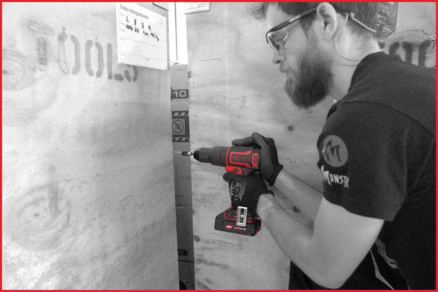 Ein Mann in Schutzbrille und Handschuhen bohrt mit einem Akkubohrer ein großes Holzbrett.