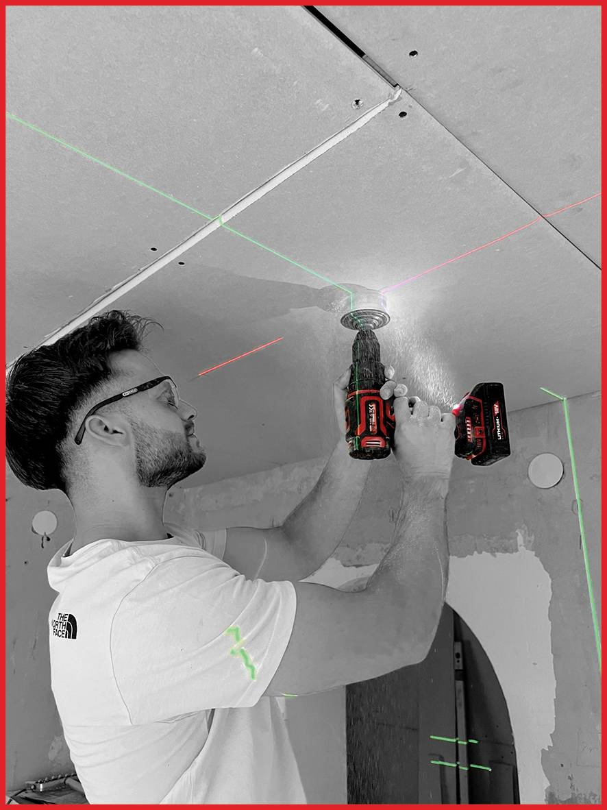 Ein Mann befestigt eine Deckenplatte mit einer Bohrmaschine. Er arbeitet in einem renovierungsbedürftigen Raum mit sichtbaren Kabeln an der Wand.