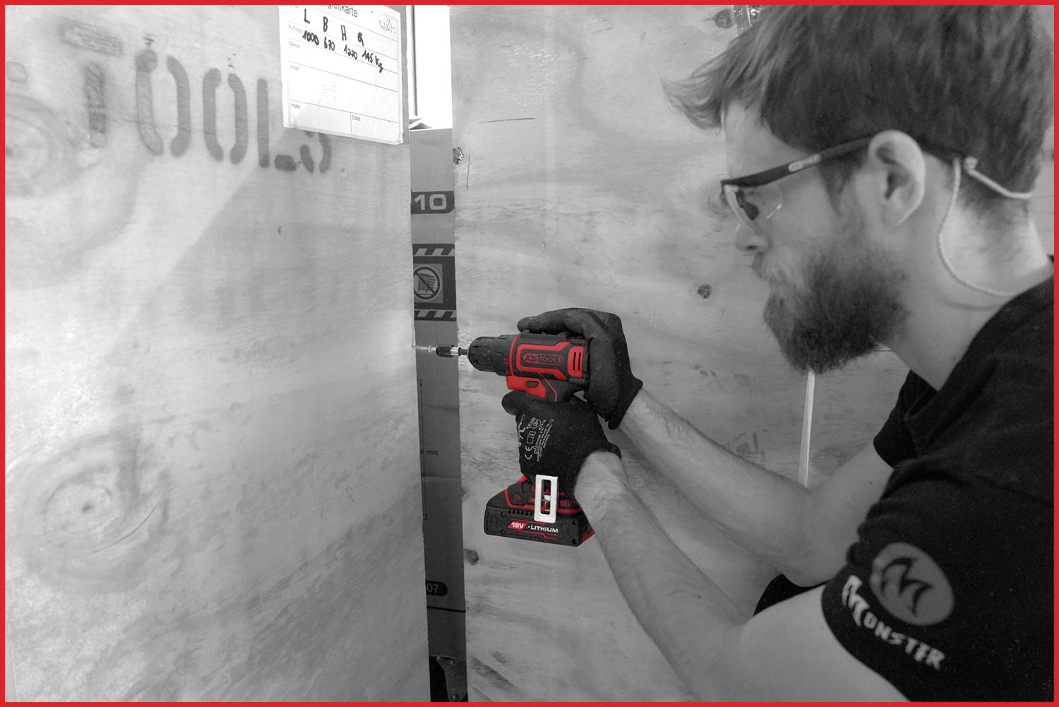 Ein Mann verwendet eine rote Bohrmaschine, um Holzplatten zusammenzubauen. Im Hintergrund sind Kartons und ein aufgehängter Zettel sichtbar.