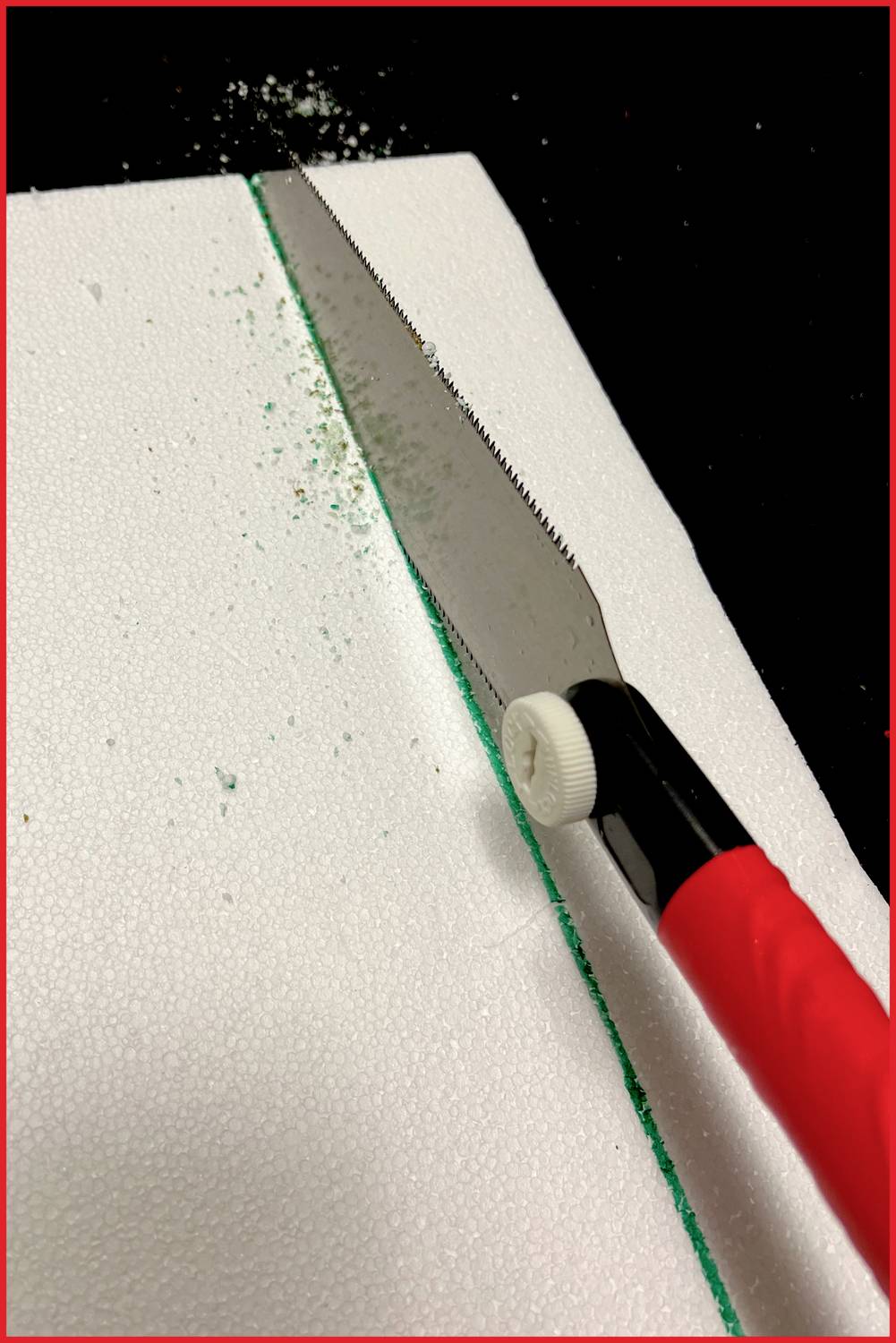 Eine rote Handsäge schneidet durch weißes Styropor. Grünen Staub sammelt sich entlang des Schnitts.