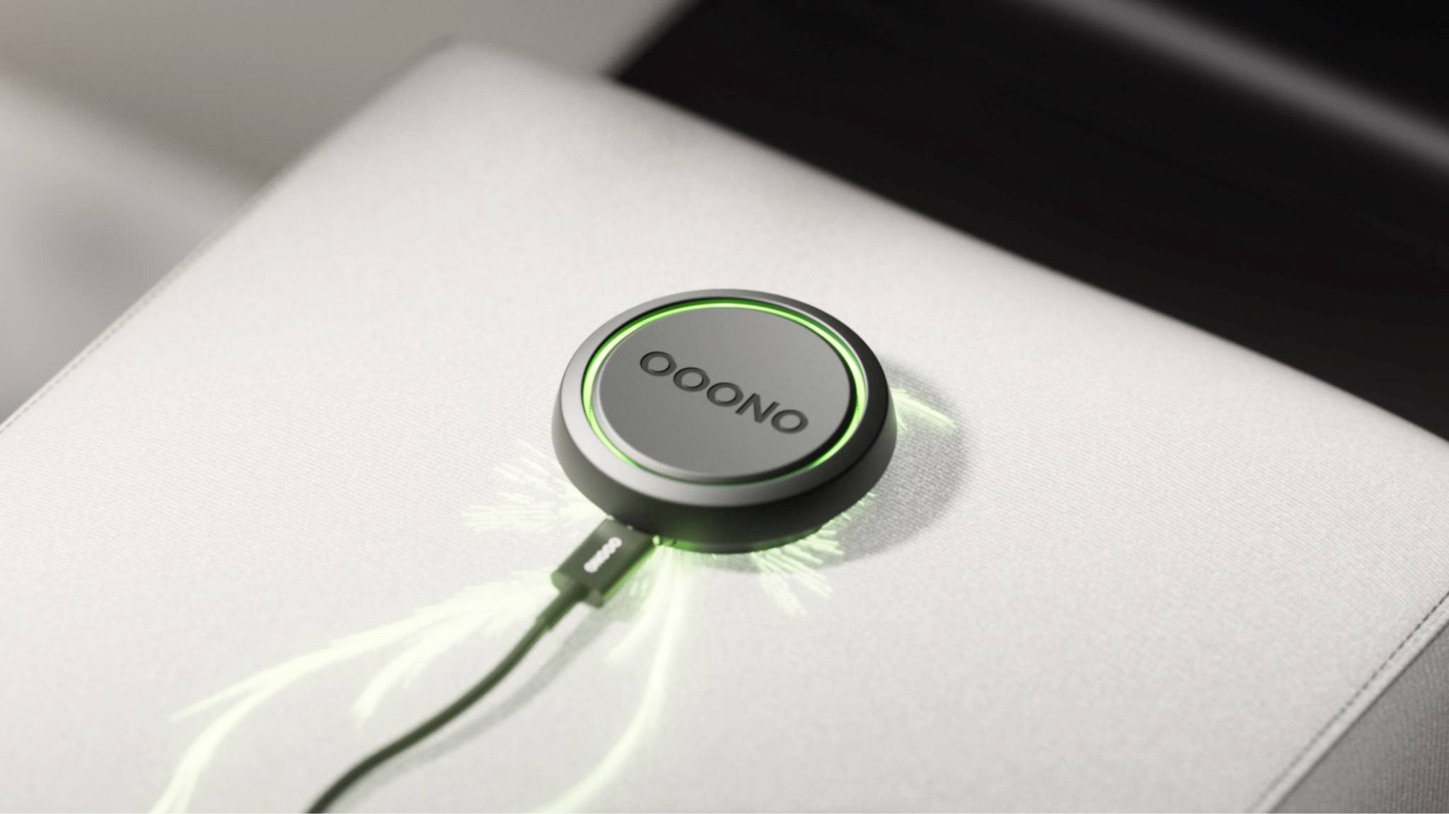 Ein rundes Gerät mit einem Kabel, auf dem 'OOONO' steht, leuchtet grün. Es liegt auf einer hellen Oberfläche.