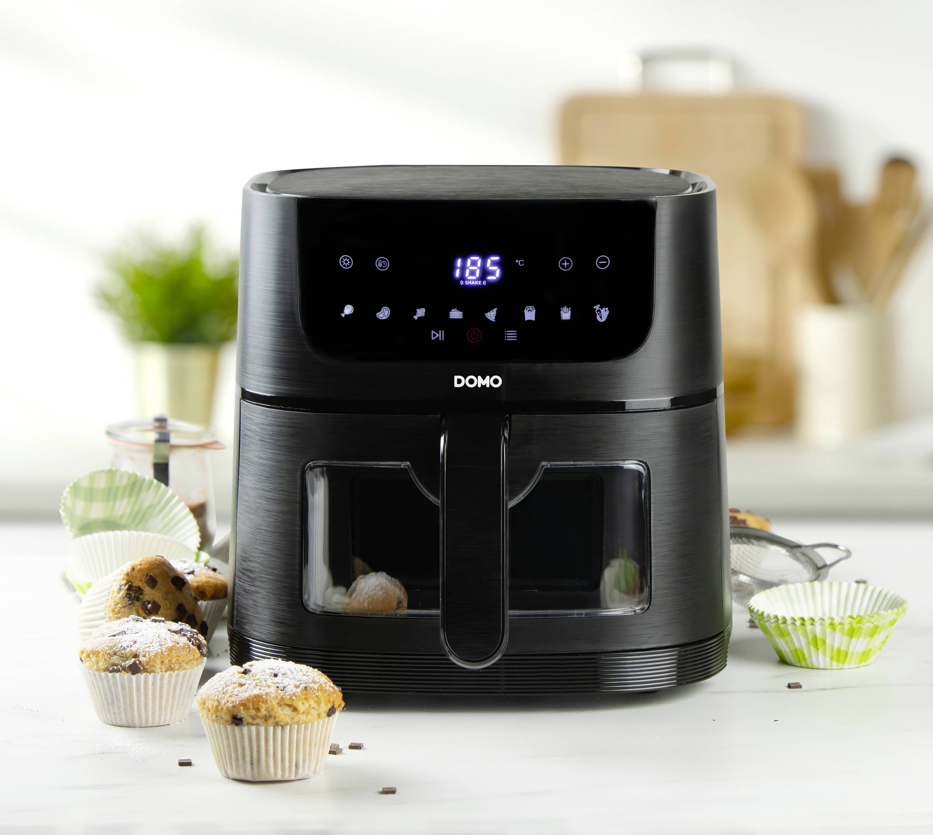 Schwarze Heißluftfritteuse auf weißem Tisch mit digitalem Display, umgeben von Muffins in bunten Förmchen, im Hintergrund Küchenutensilien.