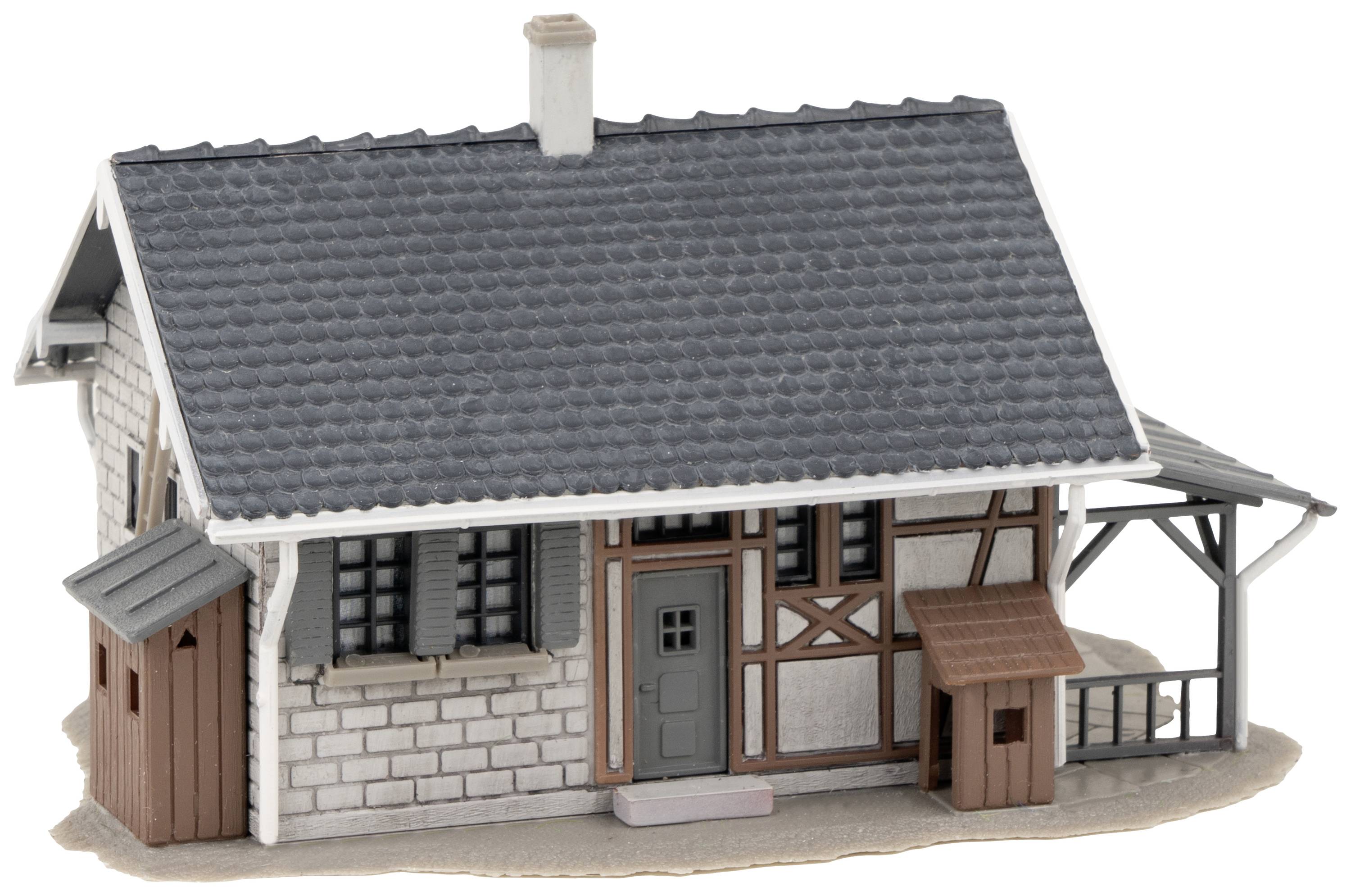 Ein kleines Modellhaus im Fachwerkstil mit grauem Dach, weißen Wänden und brauner Tür, steht auf einem sandigen Grund.