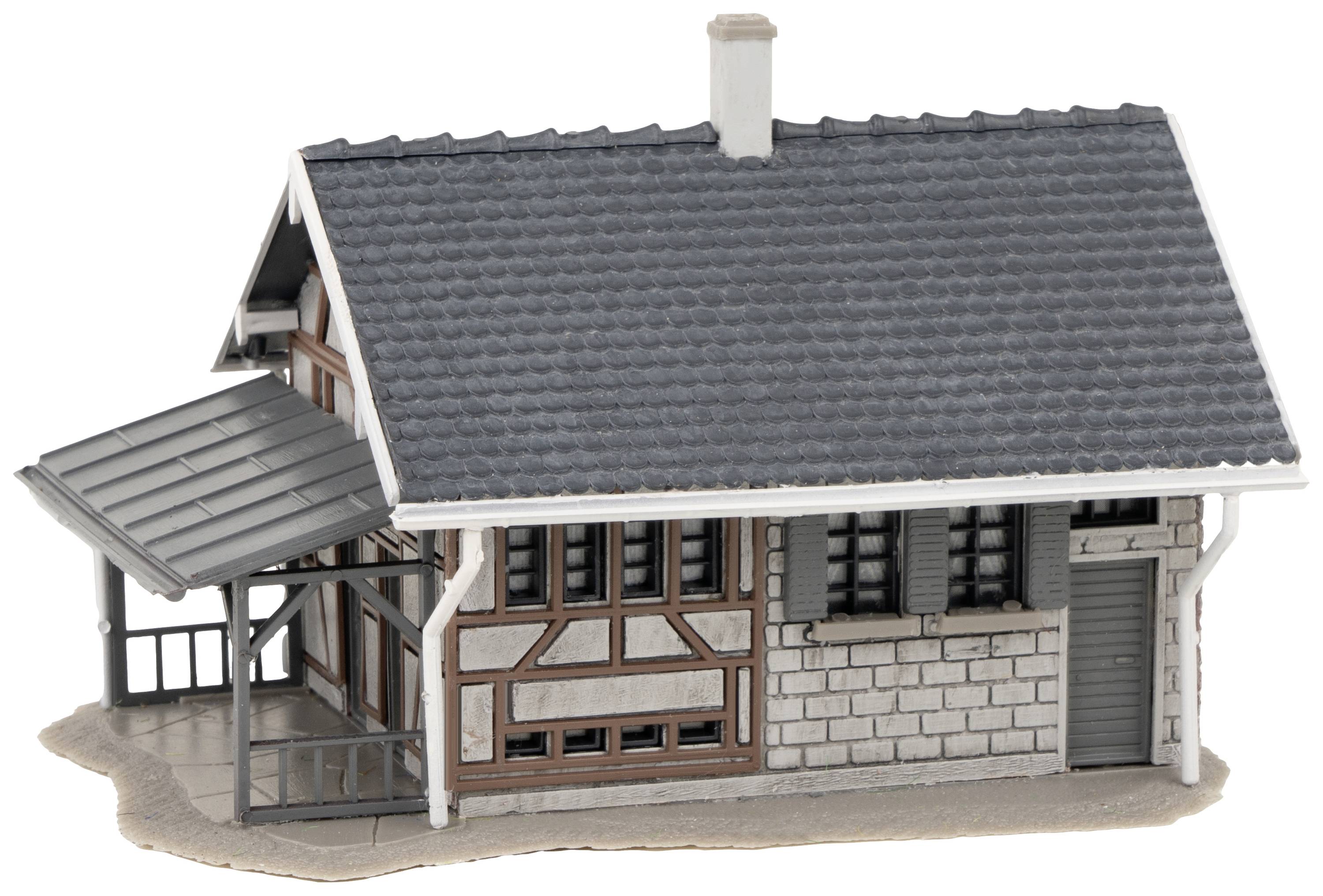 Kleines Fachwerkhausmodell mit grauem Schieferdach und angebautem Vordach. Links Holzverzierung, rechts gemauerte Wand mit Tür.
