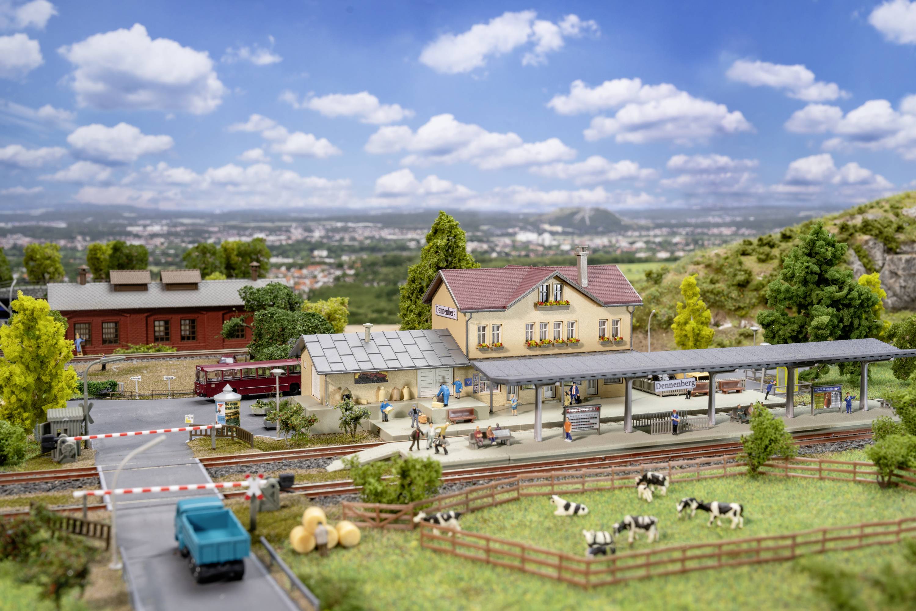 Miniatur-Modell eines ländlichen Bahnhofs mit Personen, Kühen auf einer Wiese und einer Stadt im Hintergrund.