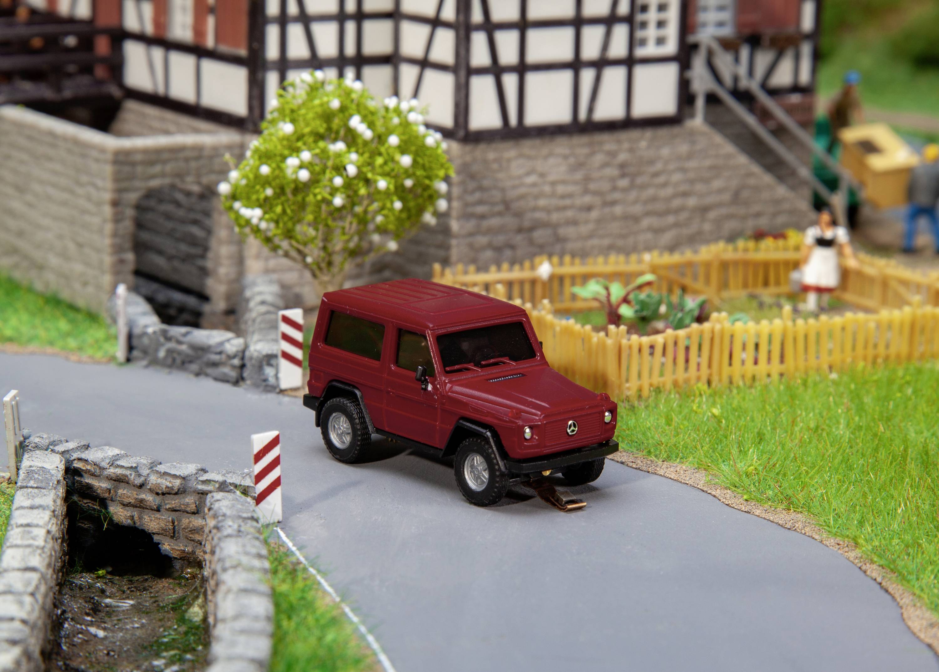 Ein rotes Modellauto parkt auf einer ländlichen Straße vor einem Fachwerkhaus mit Gartenzaun und Baum im Hintergrund.