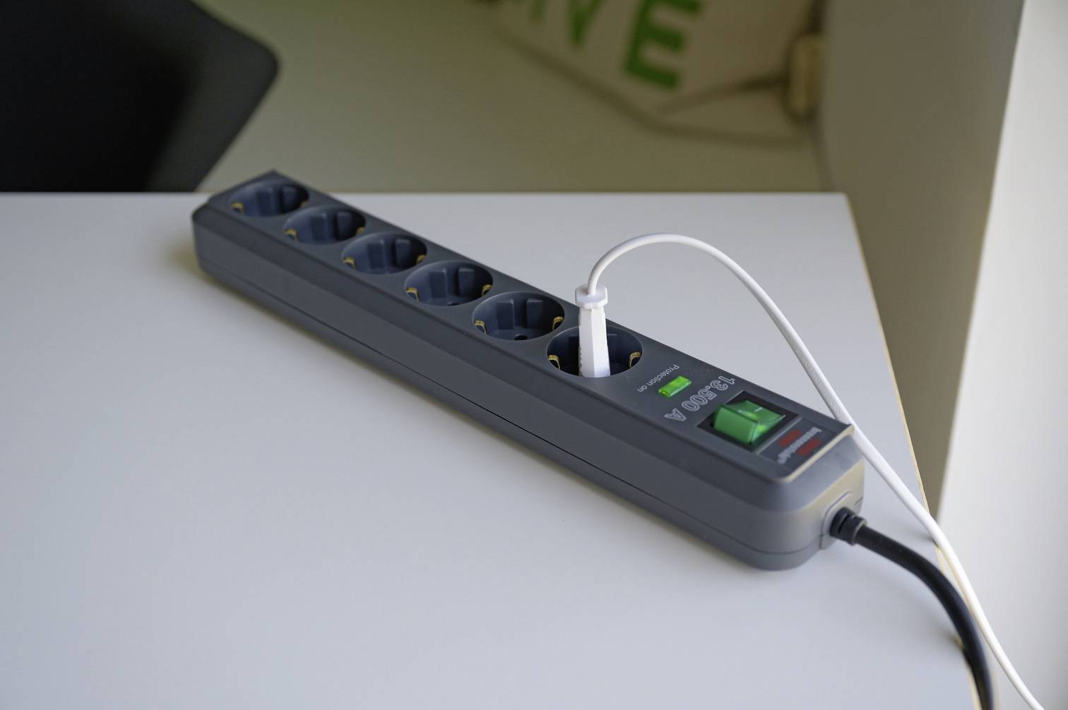 Eine graue Steckdosenleiste mit sechs Buchsen liegt auf einem Tisch. Ein weißes Ladekabel ist angeschlossen. Ein grüner Schalter ist sichtbar.