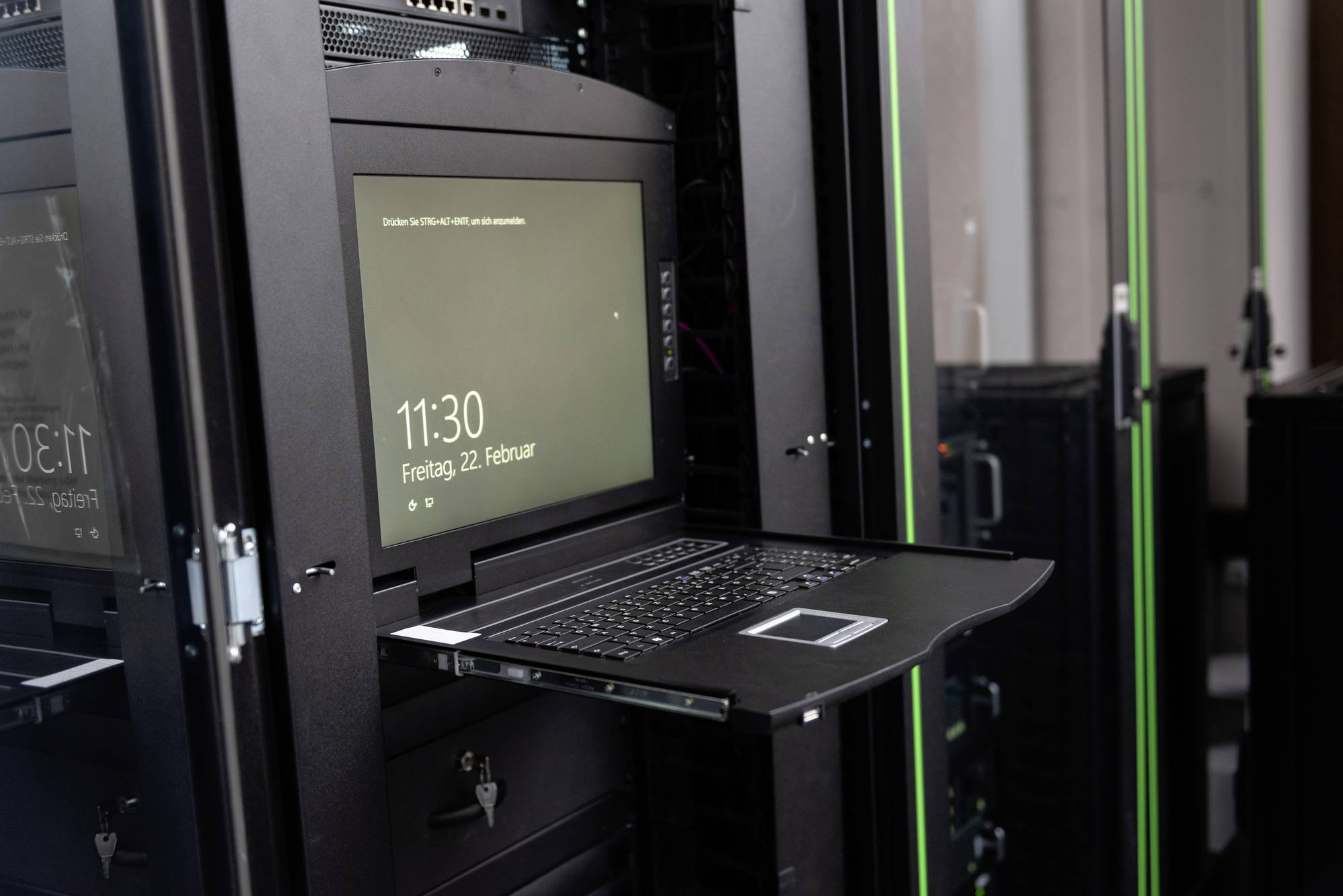 Ein Laptop in einem Serverraum, auf einem ausziehbaren Regal, zeigt Datum und Uhrzeit 'Freitag, 22 Februar, 11:30' auf dem Bildschirm.