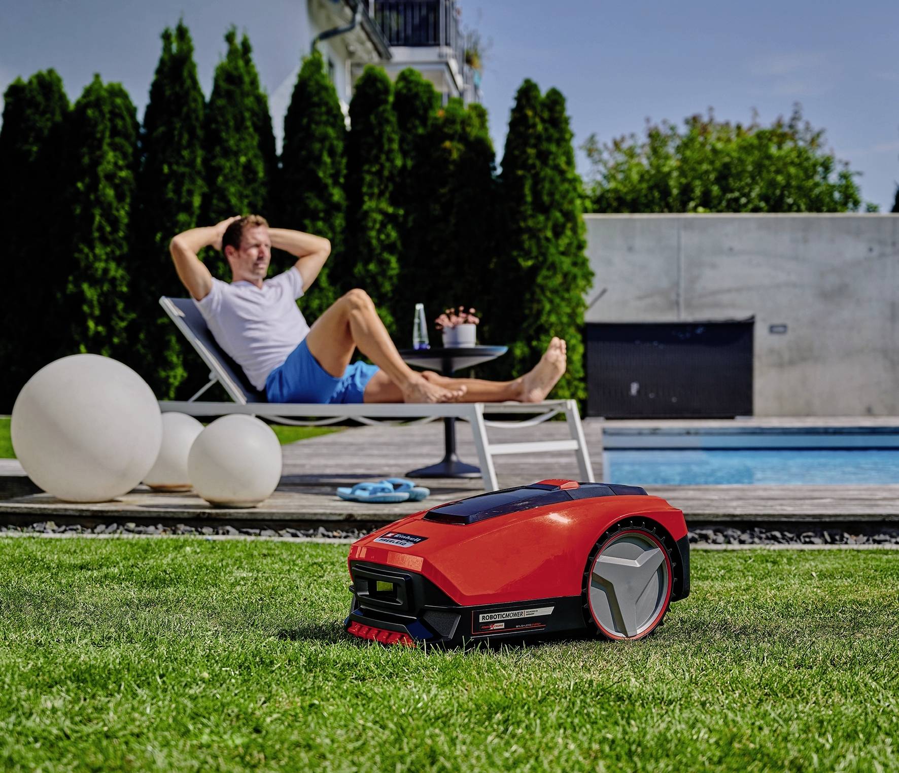 Ein Mann entspannt am Pool, während ein roter Mähroboter den Rasen mäht.