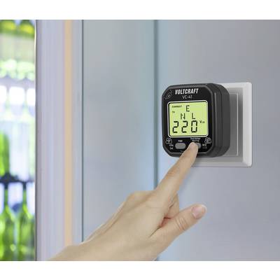 Eine Hand drückt auf ein elektrisches Messgerät mit Anzeige '220 V'. Das Gerät ist an einer Wandsteckdose angeschlossen.
