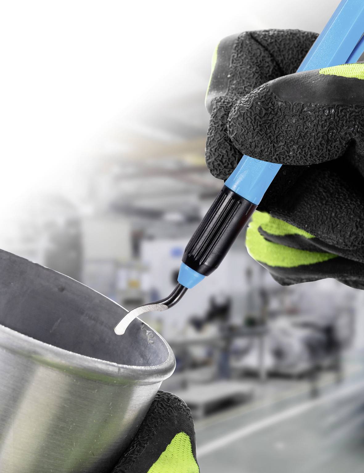 Eine Hand in Handschuh hält ein blaues Lötkolbenwerkzeug, das auf einem Metallstück arbeitet. Im Hintergrund ist eine Werkstatt unscharf zu sehen.