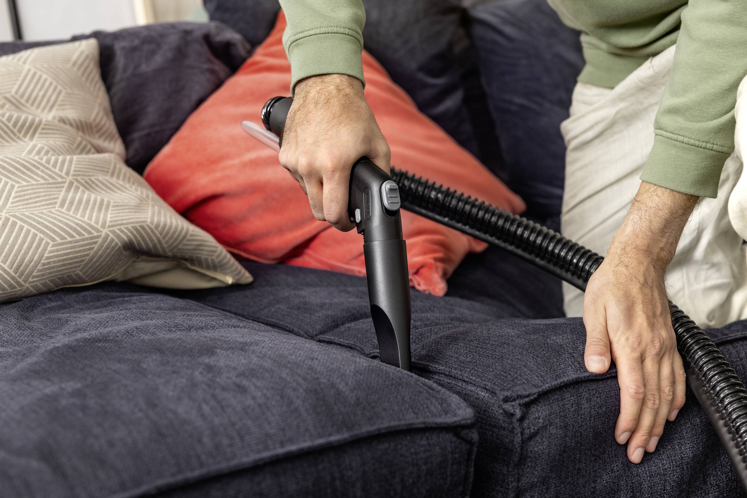 Eine Person saugt mit einem Handstaubsauger ein dunkelgraues Sofa ab. Im Hintergrund sind Kissen in verschiedenen Farben sichtbar.