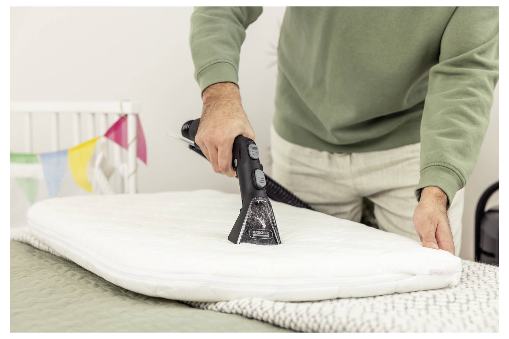 Eine Person in einem grünen Pullover dampft ein weißes Kissen auf einem Bett, während im Hintergrund bunte Flaggen hängen und auf Hausreinigung hindeuten.