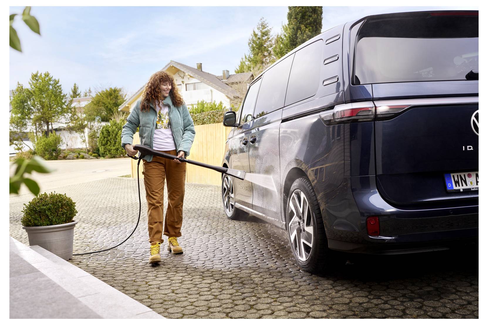 Eine Person wäscht einen dunkelblau lackierten Transporter auf einer Einfahrt und verwendet dabei einen Schlauch, der an einen Hochdruckreiniger angeschlossen ist. Daneben steht ein Blumentopf auf dem Gehweg.