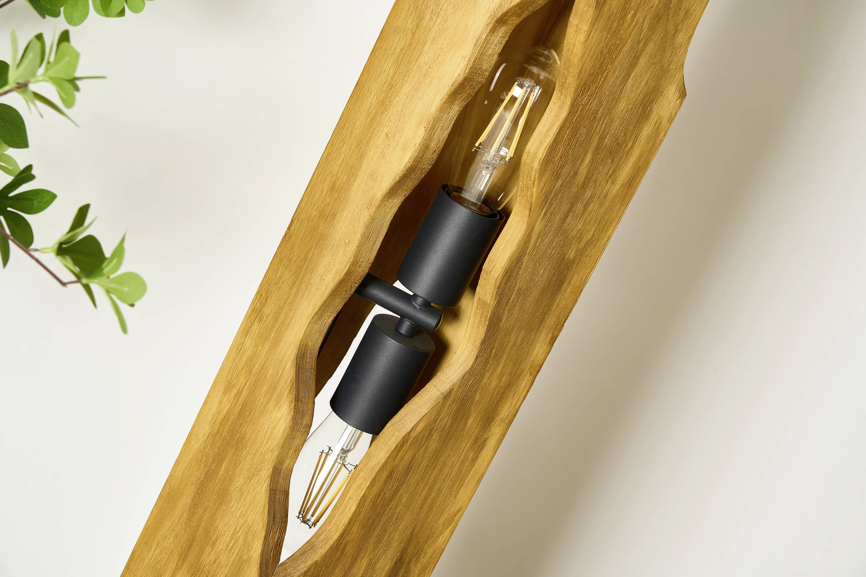 Einzigartige, handgefertigte Holzlampe mit integrierter Glühbirne, die warmes Licht abgibt. Dekorative Pflanze im Hintergrund.