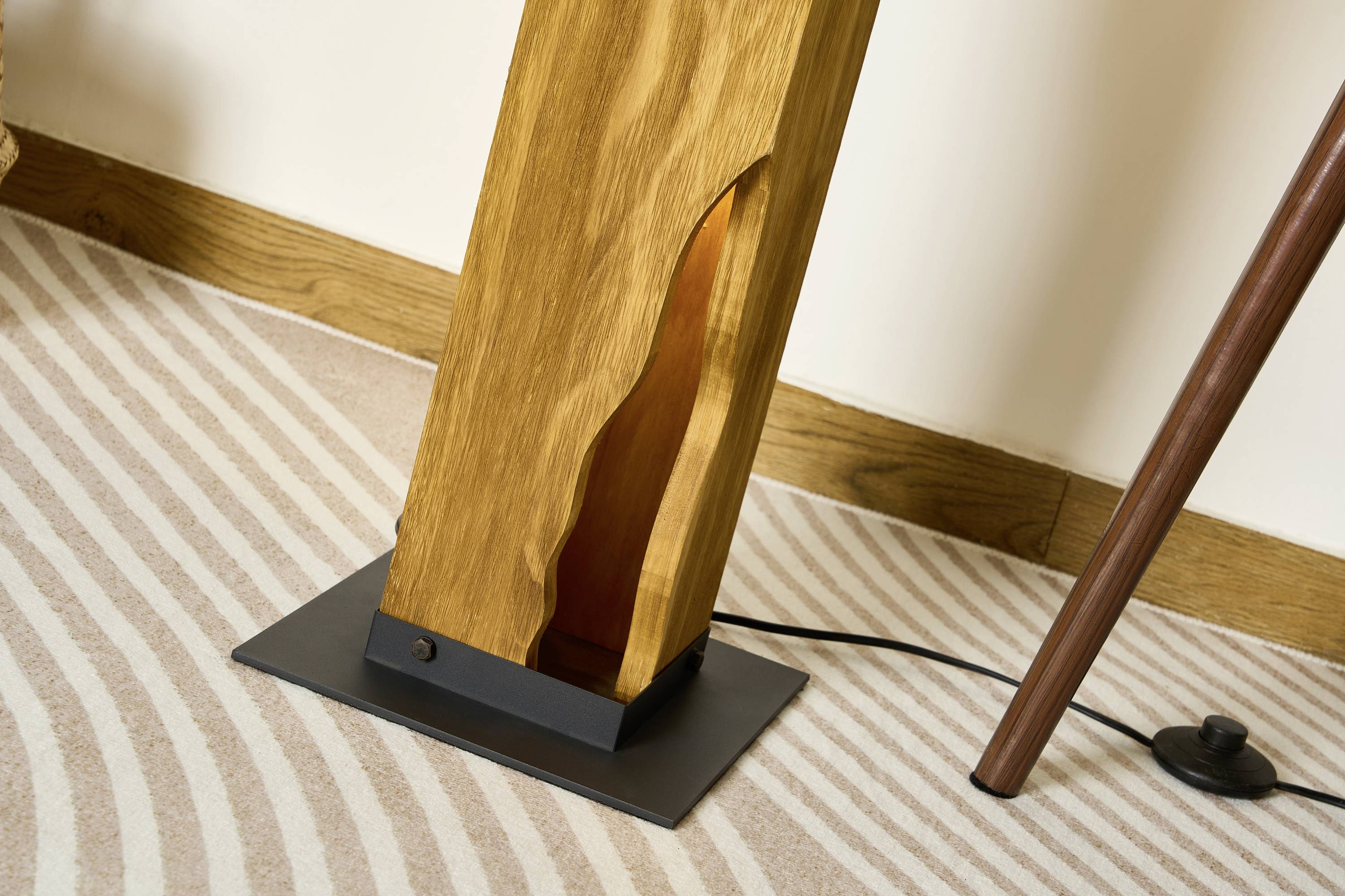 Holzlampensockel mit organischem Ausschnitt auf gestreiftem Teppich, daneben ein Holzstuhlbein. Stromkabel sichtbar.