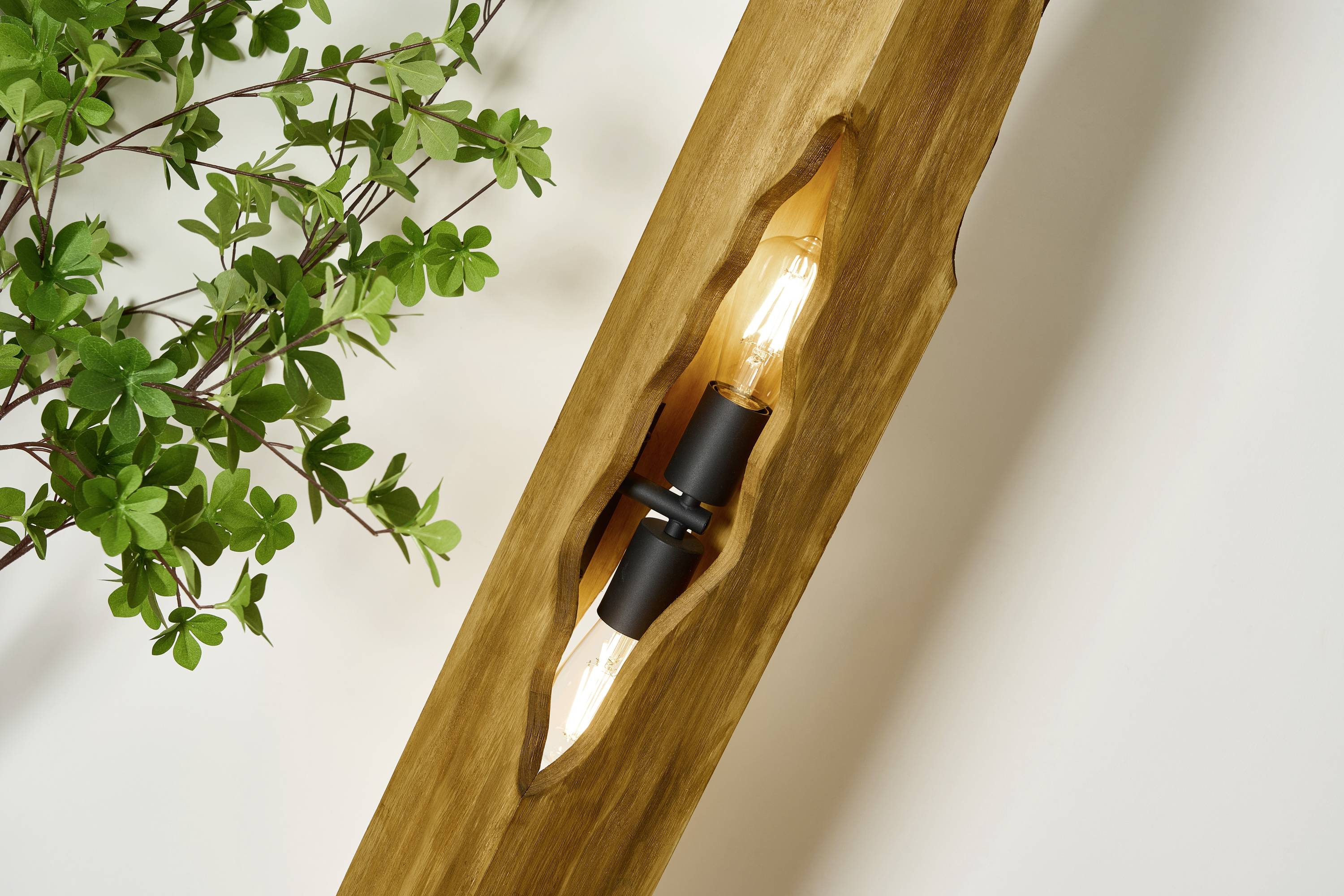 Eine moderne Wandlampe aus Holz mit sichtbare Glühbirne, neben einem Zweig grüner Blätter an einer weißen Wand.
