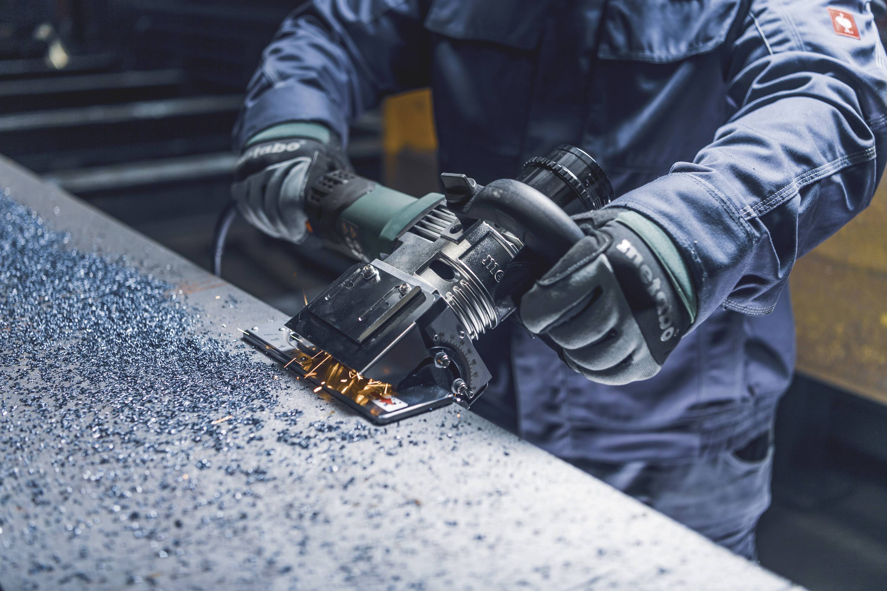 Arbeiter schleift Metallplatte mit Elektrowerkzeug, Metallspäne verstreut. Trägt blaue Schutzkleidung und Handschuhe.