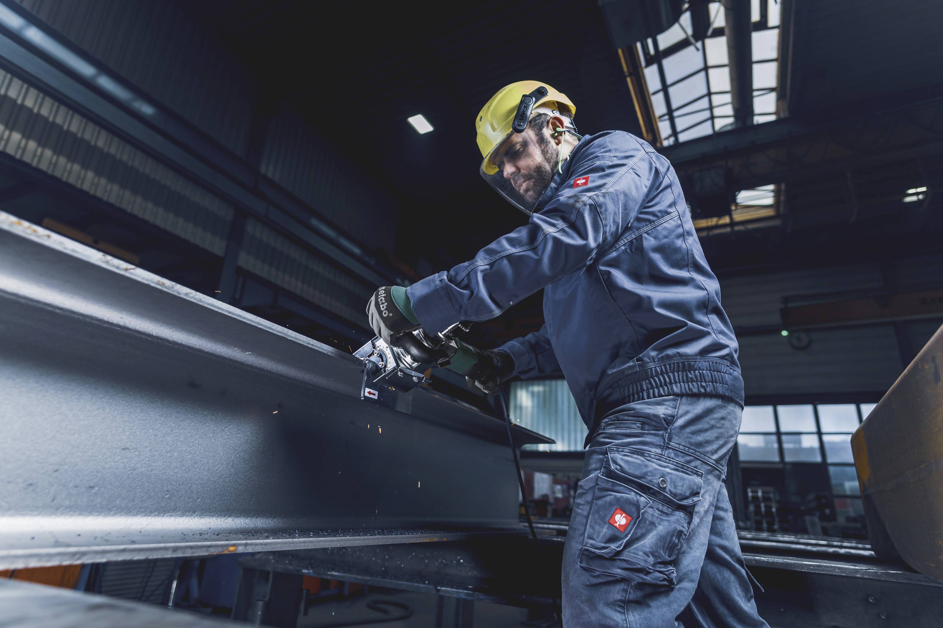Ein Arbeiter in Schutzkleidung schleift mit einem Werkzeug eine Metallträger. Die Umgebung ist eine industrielle Werkhalle.