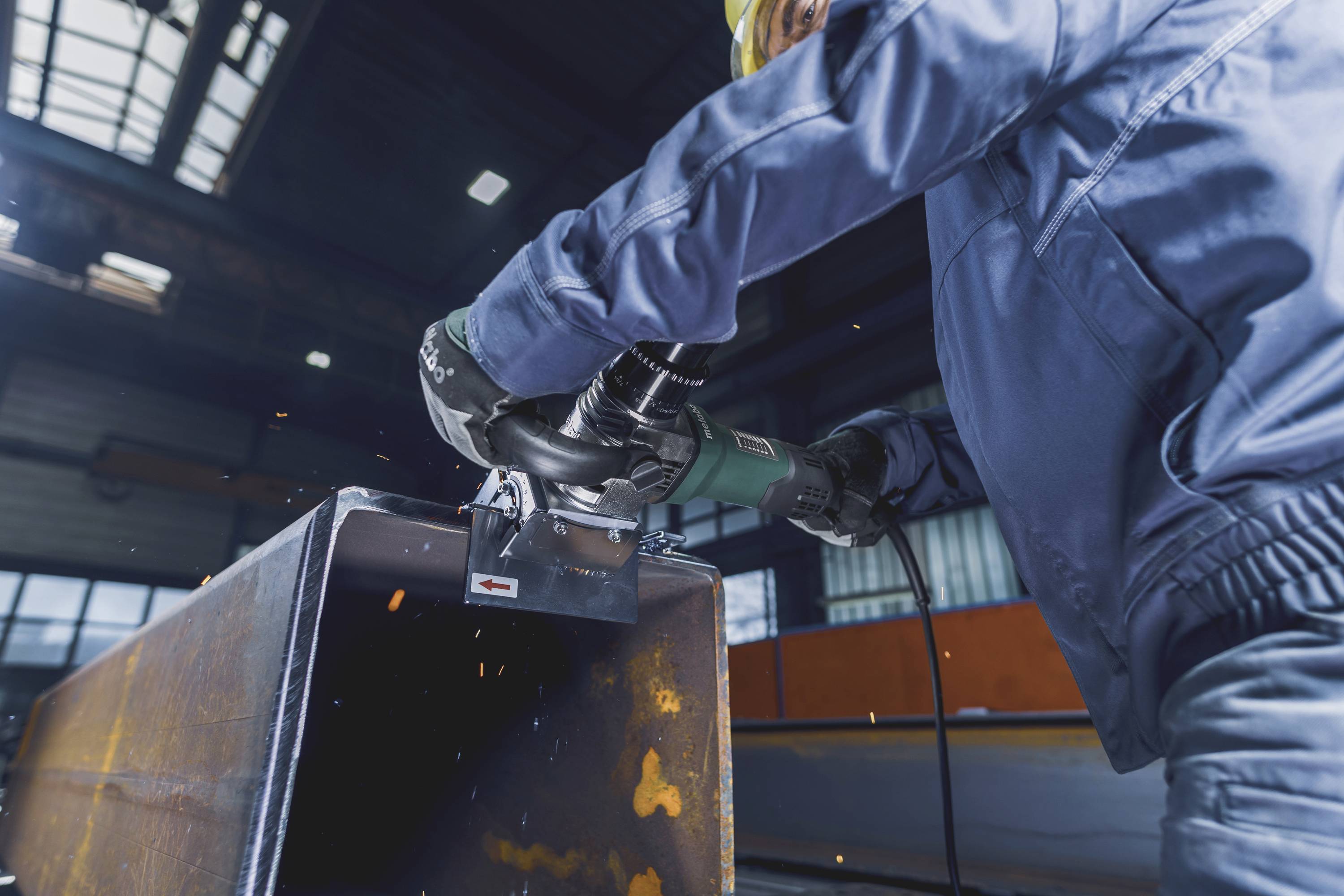 Eine Person schleift Metall in einer Werkstatt. Funken fliegen vom Werkstück, während die Person Schutzkleidung trägt.