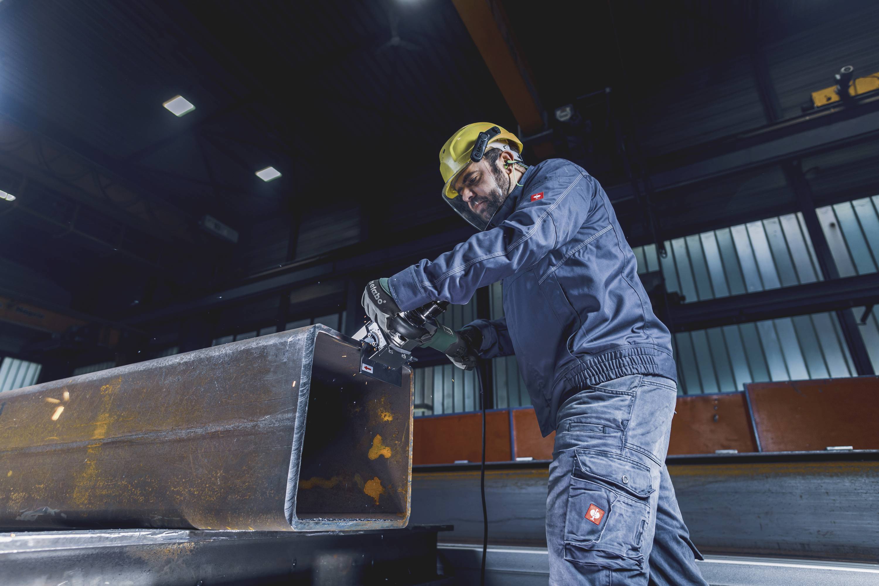 Ein Arbeiter mit Helm und Schutzkleidung schleift ein großes Metallstück in einer Fabrikhalle. Die Szene wirkt industrienahe und funktional.