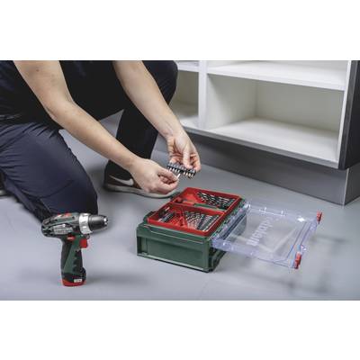 Person sortiert Bohrer in einem grünen Werkzeugkasten neben einer Bohrmaschine auf dem Boden vor einem weißen Regal.