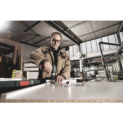 Ein Mann arbeitet in einer Werkstatt und benutzt eine Bohrmaschine, um auf einer Werkbank zu arbeiten. Im Hintergrund sind Maschinen sichtbar.