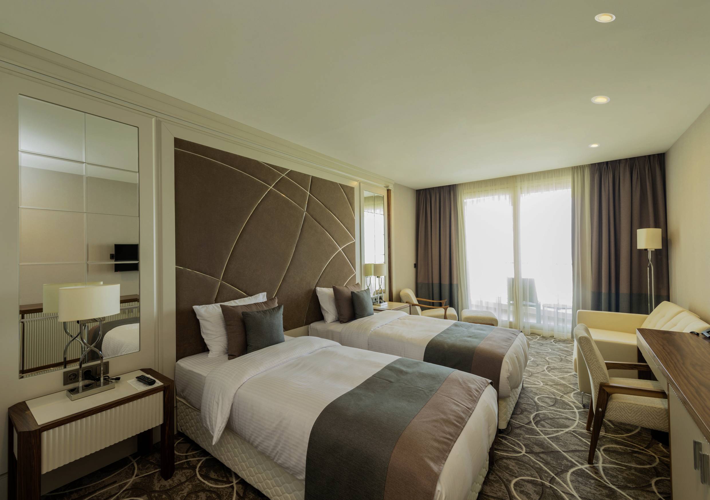 Ein modernes Hotelzimmer mit zwei Einzelbetten, einem Schreibtisch, zwei Stühlen und großen Fenstern mit Vorhängen, die Tageslicht einlassen.