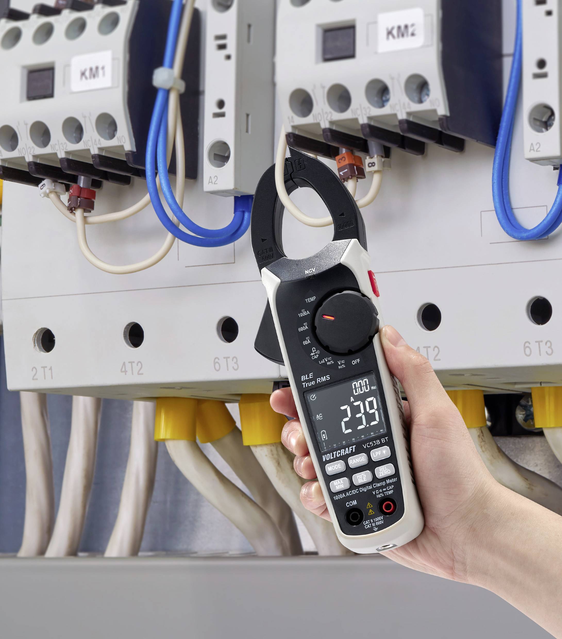 Eine Hand hält ein digitales Strommessgerät mit Zangenfunktion vor einem Schaltschrank, zeigt eine Messung von 239 Volt an.