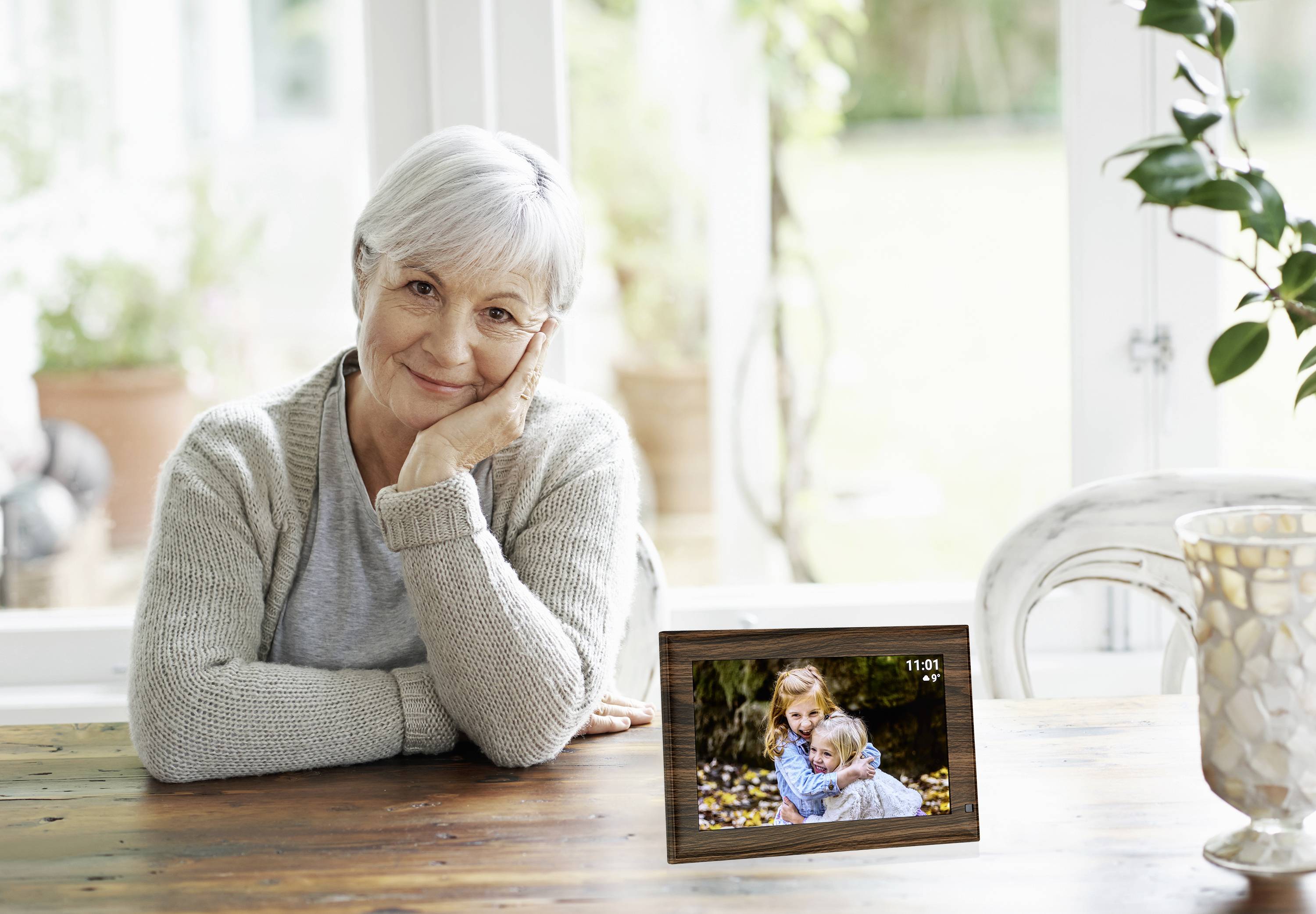 Eine ältere Frau sitzt lächelnd am Tisch, neben ihr steht ein digitaler Bilderrahmen mit einem Bild von einem Kind, das sie umarmt.