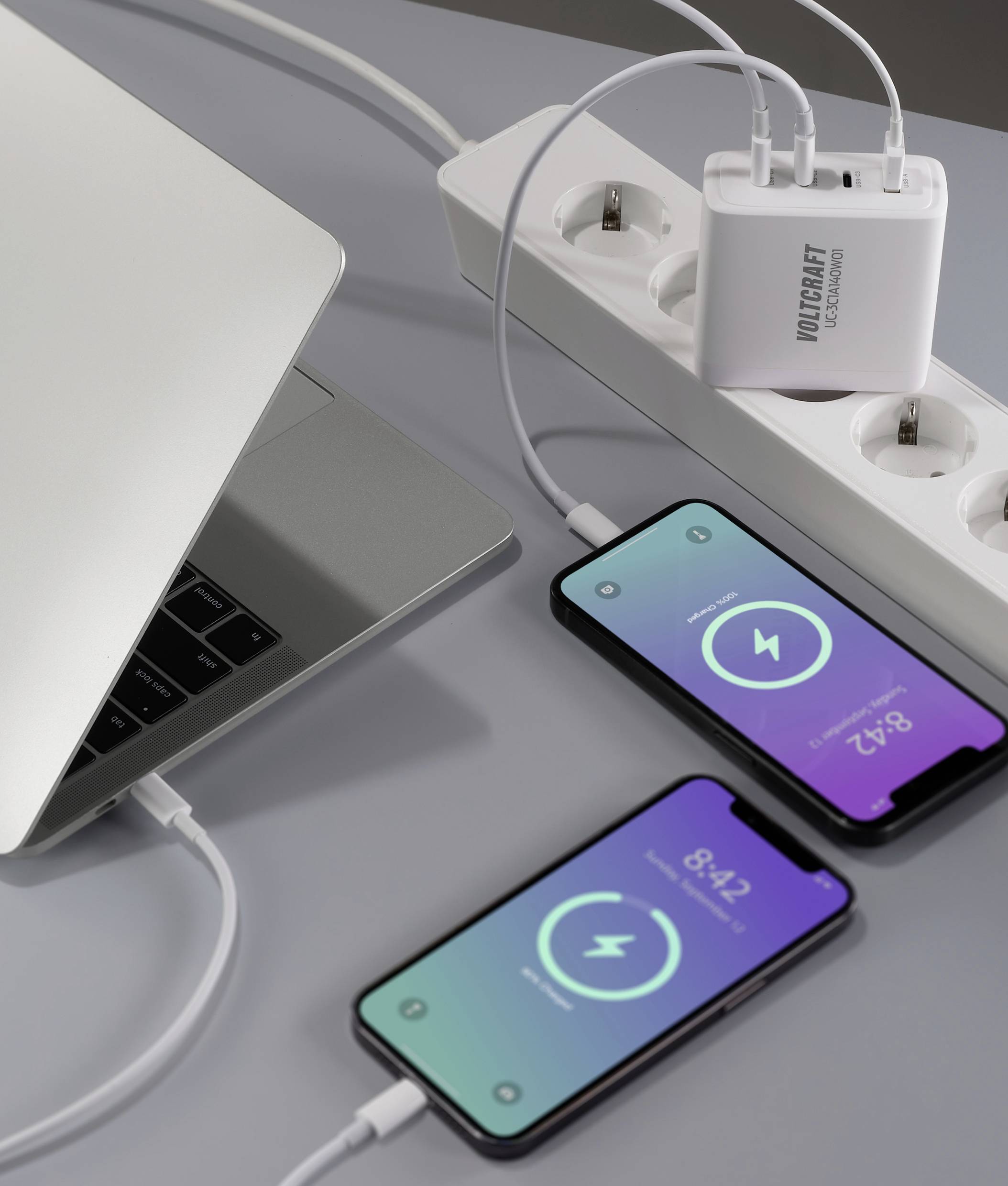 Ein Laptop und zwei Smartphones werden über ein Ladegerät an eine Steckdosenleiste angeschlossen. Die Smartphones zeigen Ladesymbole.