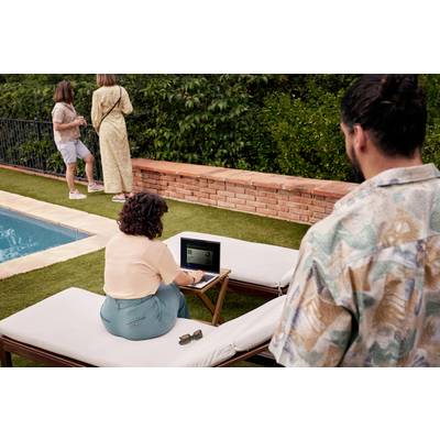 Eine Person sitzt auf einer Liege im Garten und arbeitet an einem Laptop. Zwei weitere Personen stehen am Poolrand.