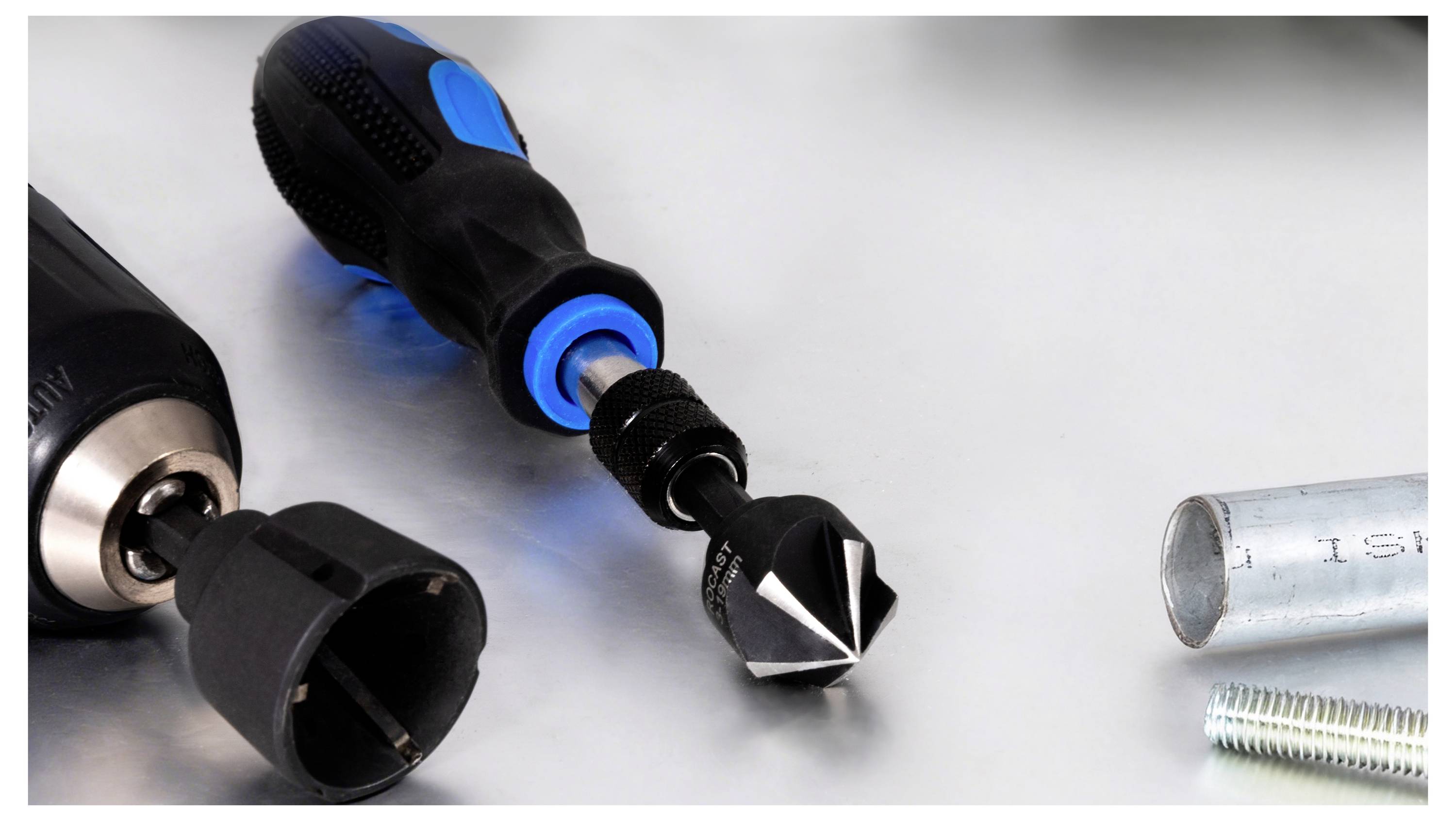 Nahaufnahme eines Schraubenziehers mit austauschbaren Bits und Steckaufsätzen, zusammen mit einer Schraube und einem Metallrohr auf einer weißen Oberfläche.