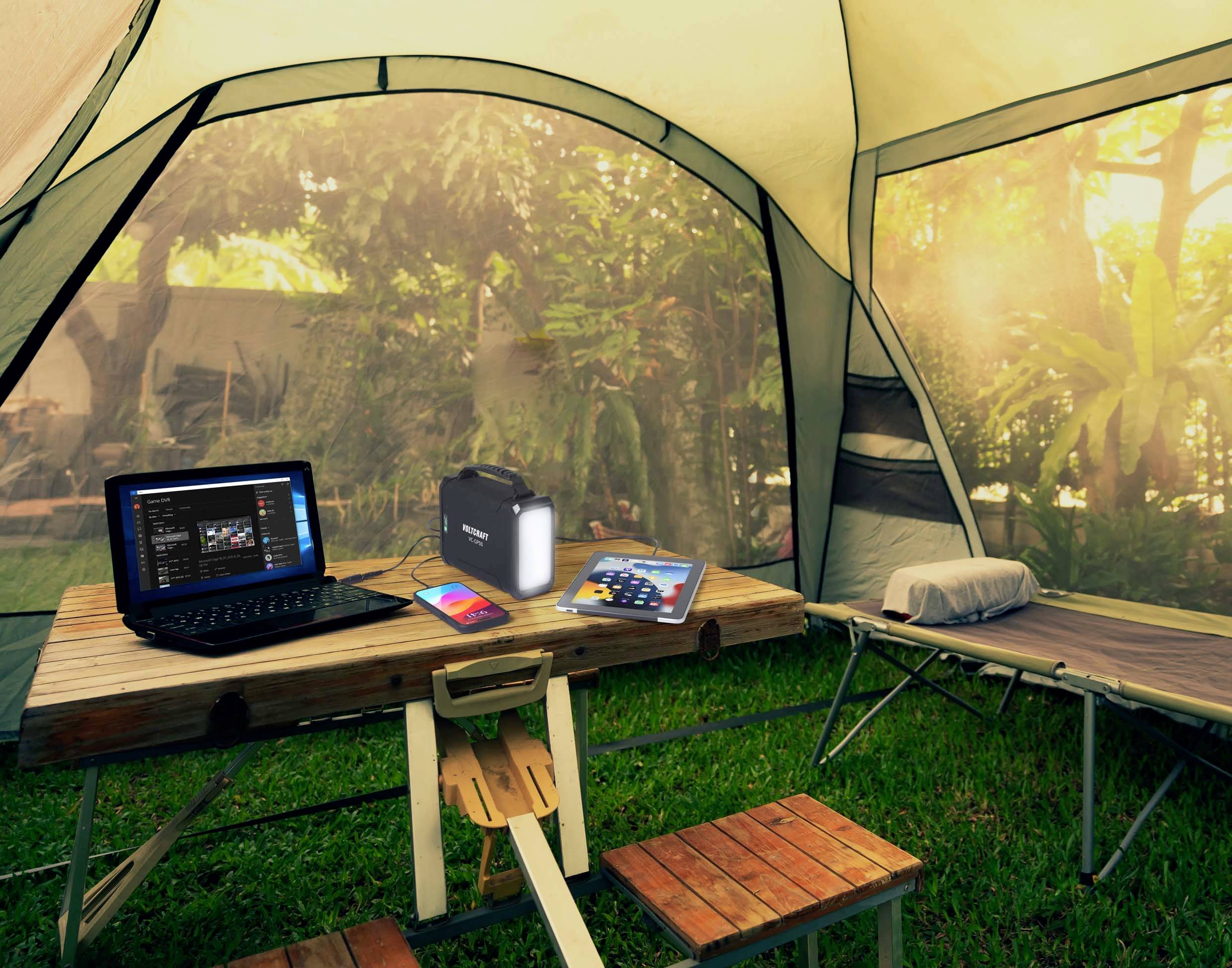 Ein Laptop, eine Powerbank und ein Tablet liegen auf einem Tisch in einem Zelt. Im Hintergrund sind Bäume. Es entsteht eine natürliche, entspannte Atmosphäre.