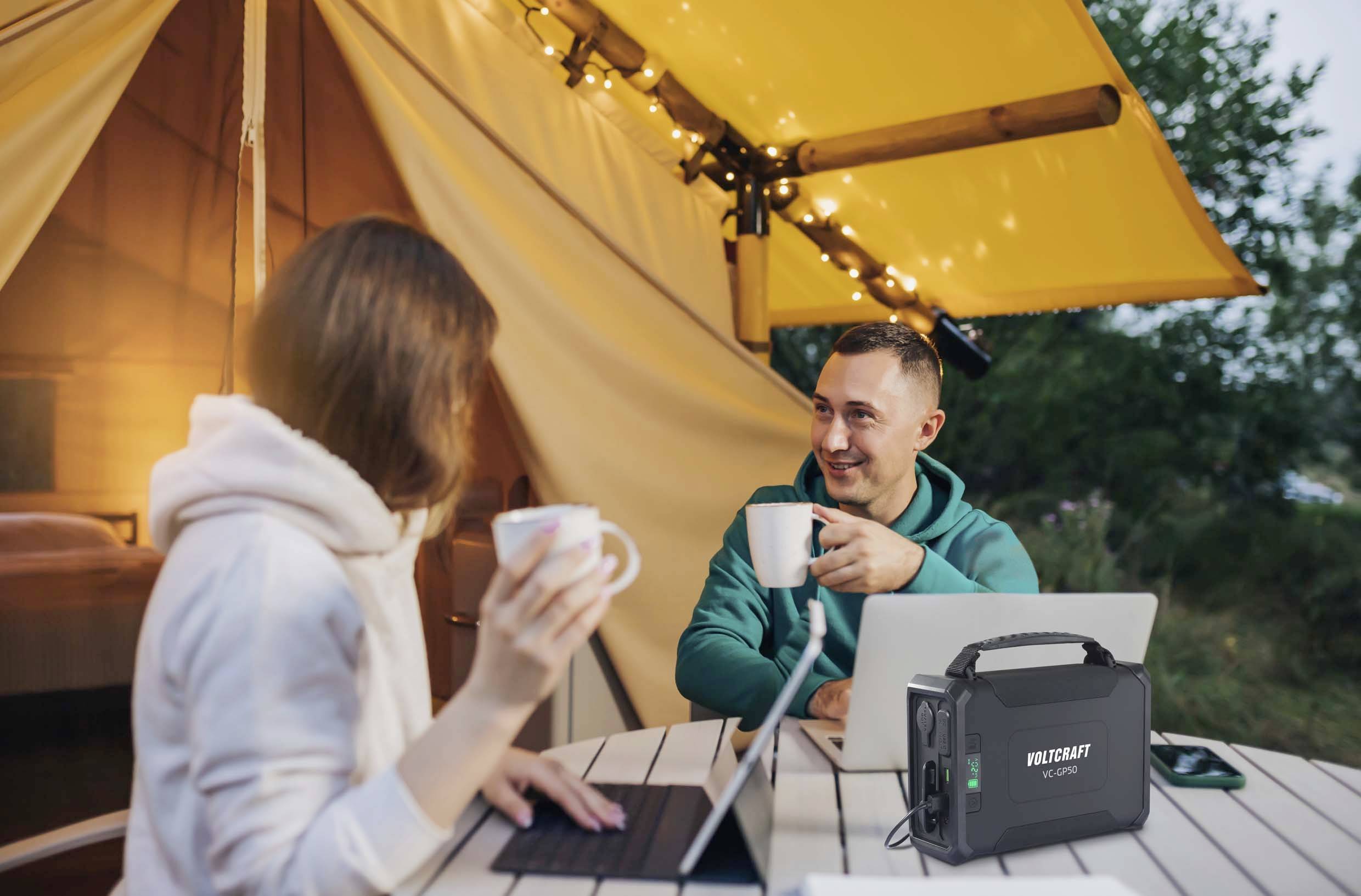 Ein Mann und eine Frau sitzen in einem Zelt unter einem gelben Vordach, trinken Kaffee und arbeiten an Laptops. Ein tragbares Stromgerät steht auf dem Tisch.