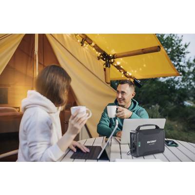 Ein Mann und eine Frau sitzen in einem Zelt unter einem gelben Vordach, trinken Kaffee und arbeiten an Laptops. Ein tragbares Stromgerät steht auf dem Tisch.
