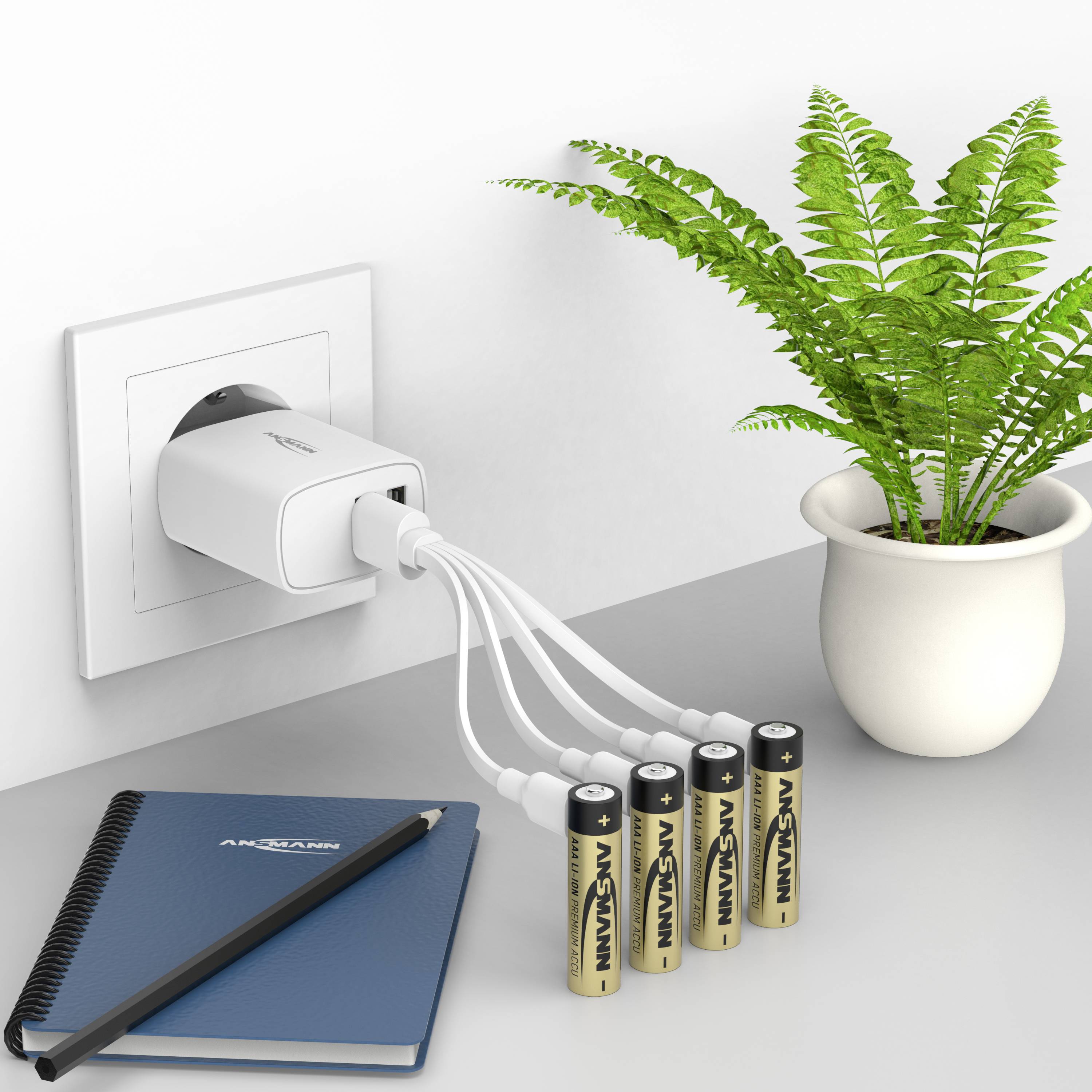 Ein Ladegerät ist in eine Steckdose eingesteckt und lädt vier AA-Batterien, die auf einem Tisch neben einem Notizbuch und einer Pflanze liegen.