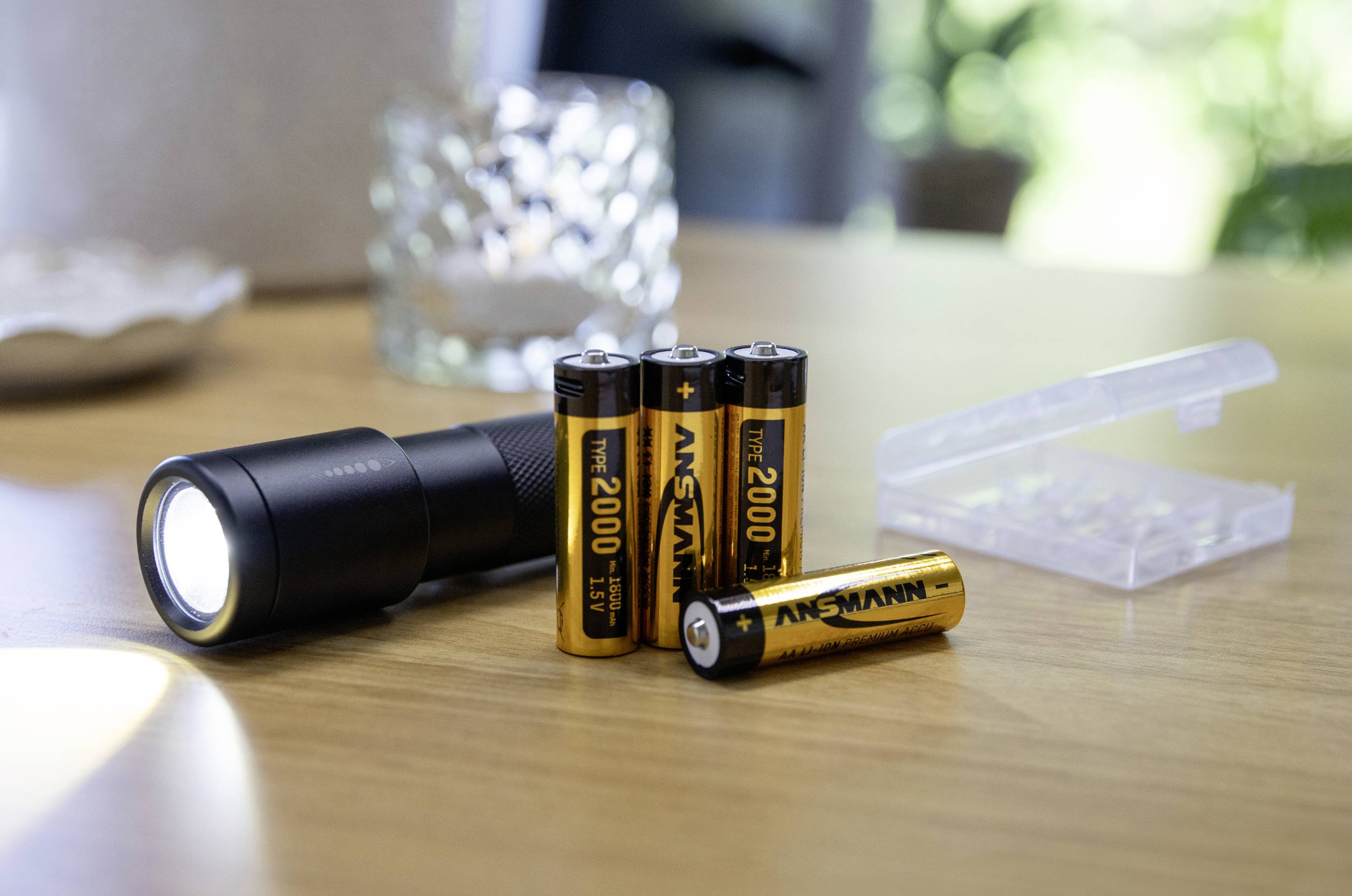 Eine schwarze Taschenlampe leuchtet auf einem Holztisch neben drei AA-Batterien und einer durchsichtigen Batteriehülle.