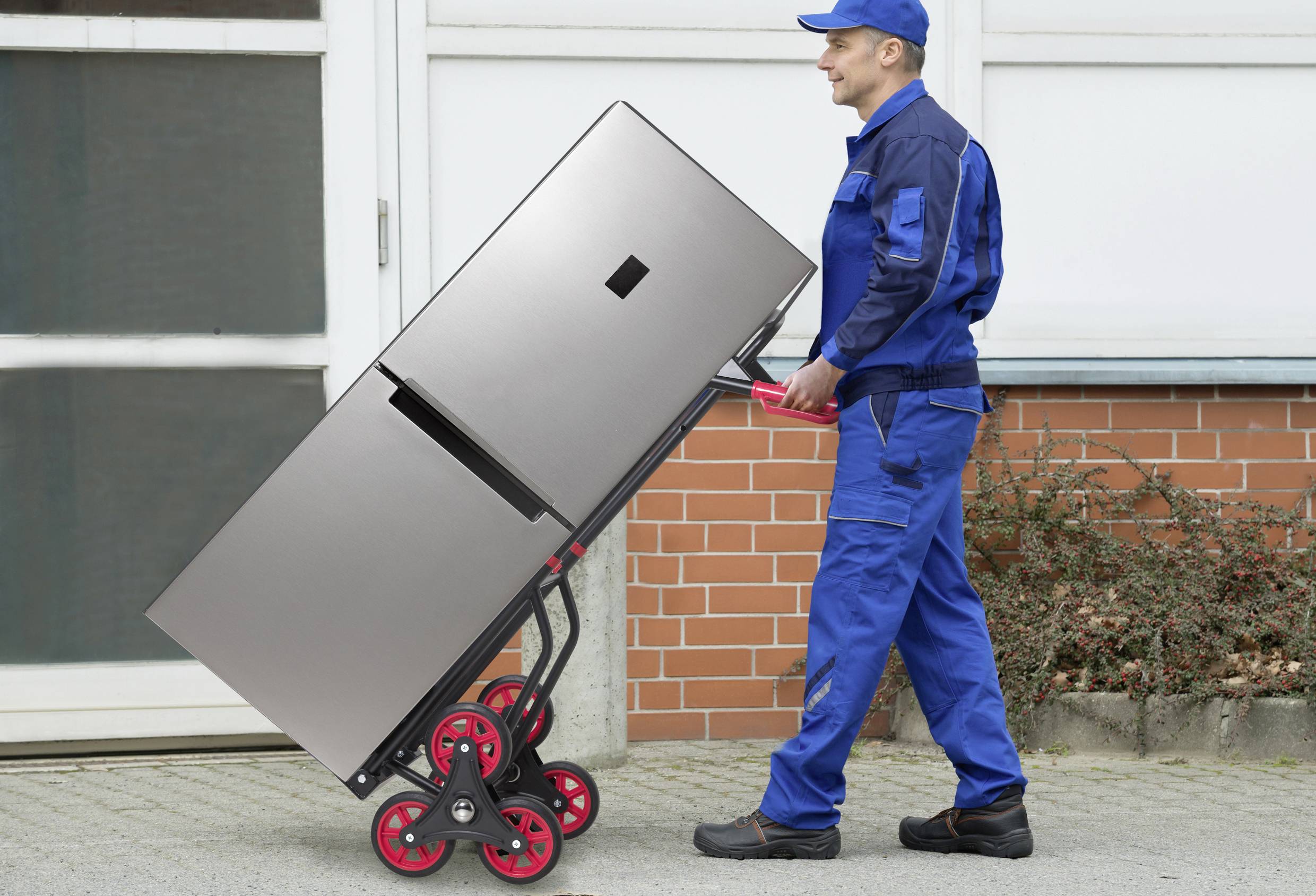 Ein Mann in blauer Uniform transportiert mit einem speziellen Rollwagen einen großen Kühlschrank vor einem Gebäude.